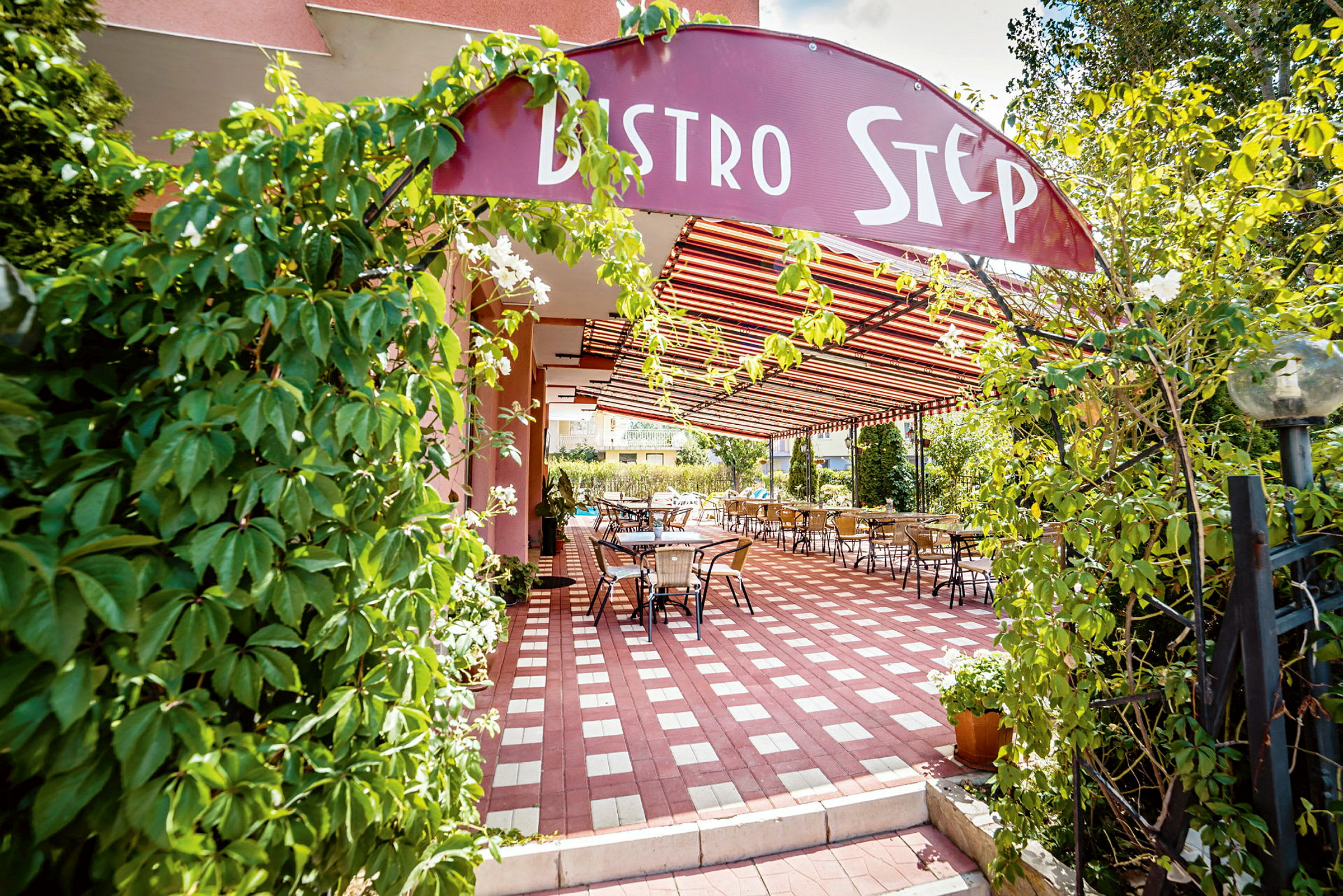 Indgang til en restaurant med et skilt "Bistro Step", omgivet af grøn beplantning og udendørs siddepladser under tag. (AI-genereret tekst)