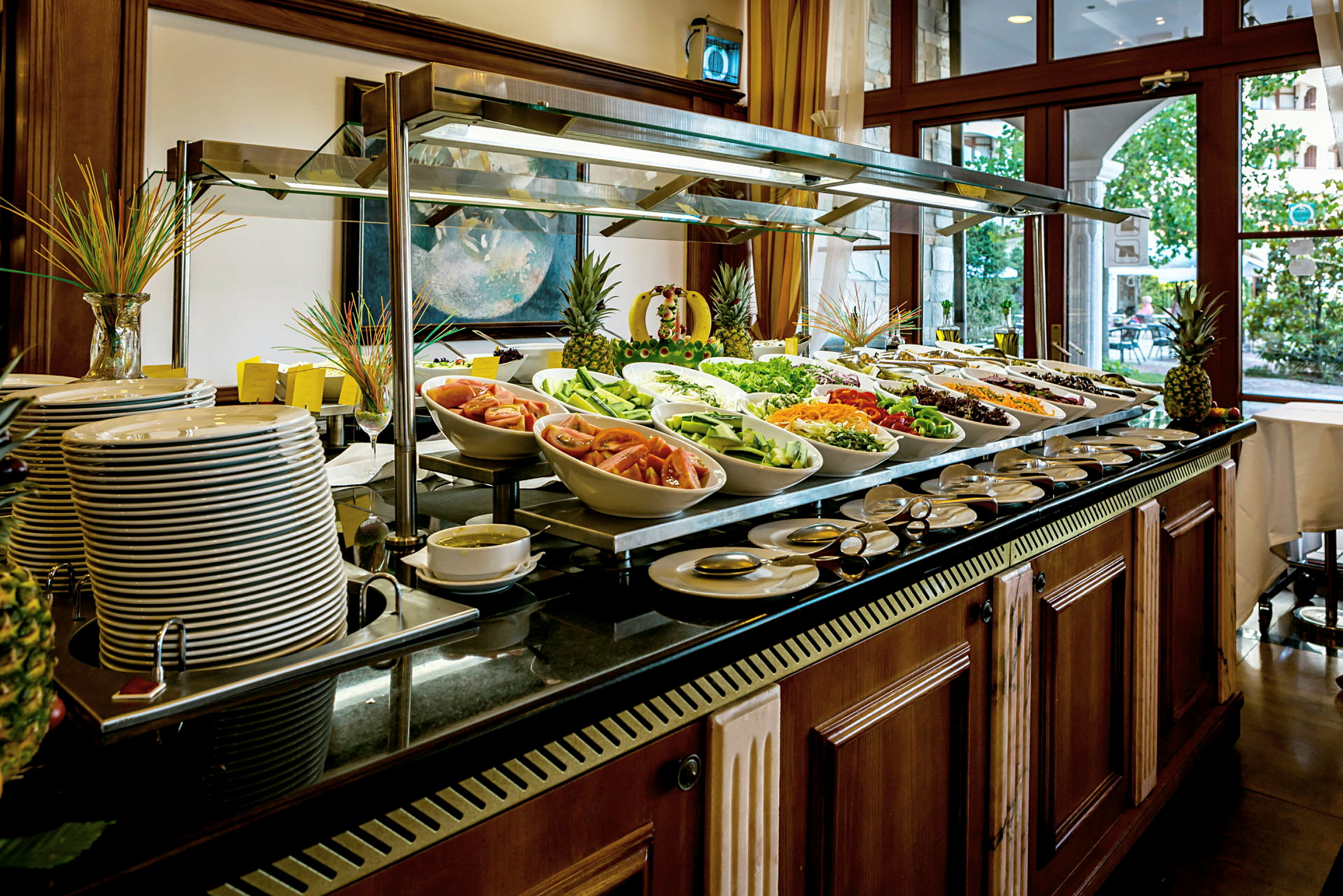 En buffet med blandt andet grøntsager, pastaretter og tallerkener. Bagved ses lysindfald fra vinduer. (AI-genereret tekst)