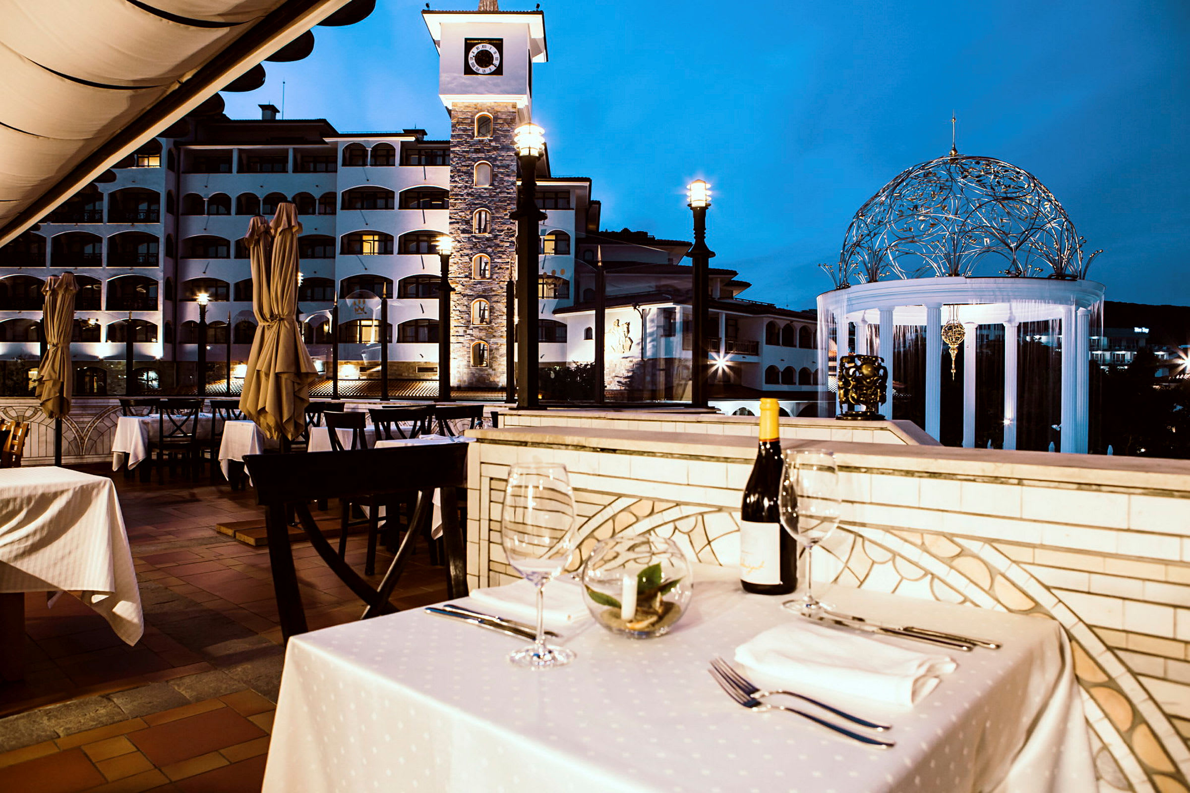 En udendørs restaurant med dækkede borde og udsigt over en bygning med et klokketårn og en oplyst pavillon. (AI-genereret tekst)