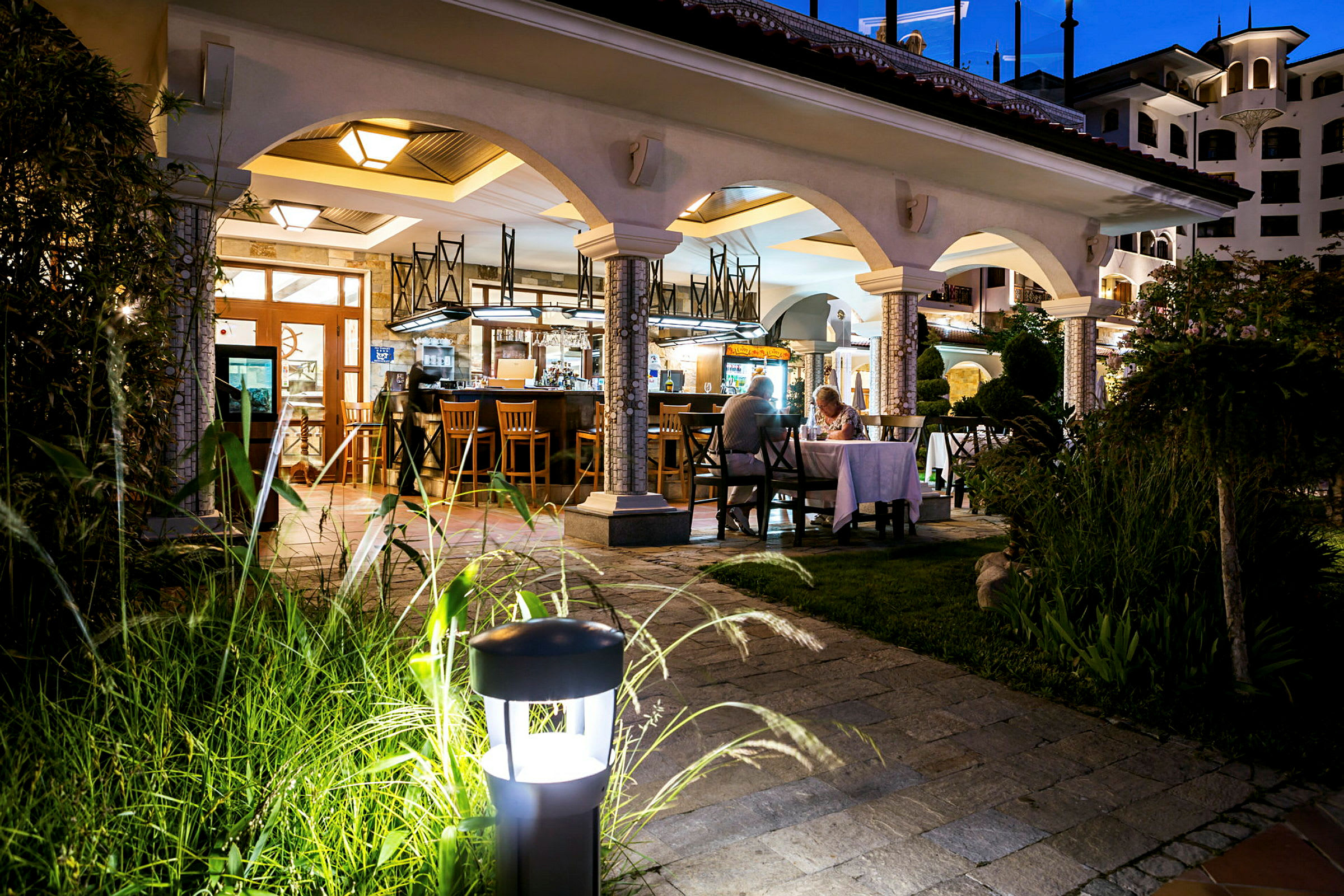 Billede af en restaurant med buet arkitektur, udendørs belysning og en elegant atmosfære om aftenen. (AI-genereret tekst)