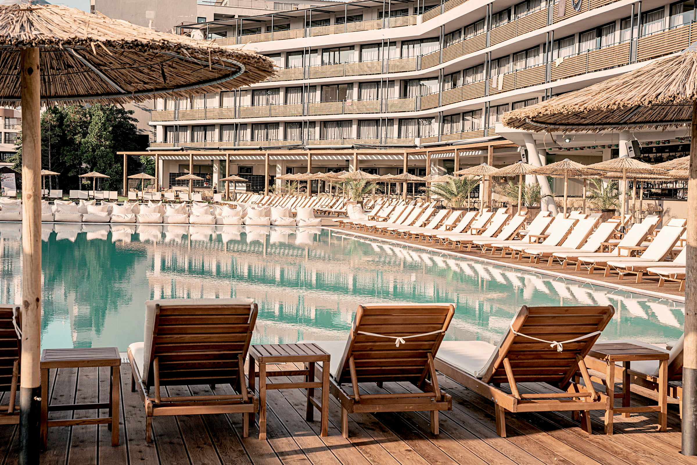 Stole og borde står ved en pool, omgivet af parasoller og et hotel i baggrunden. (AI-genereret tekst)