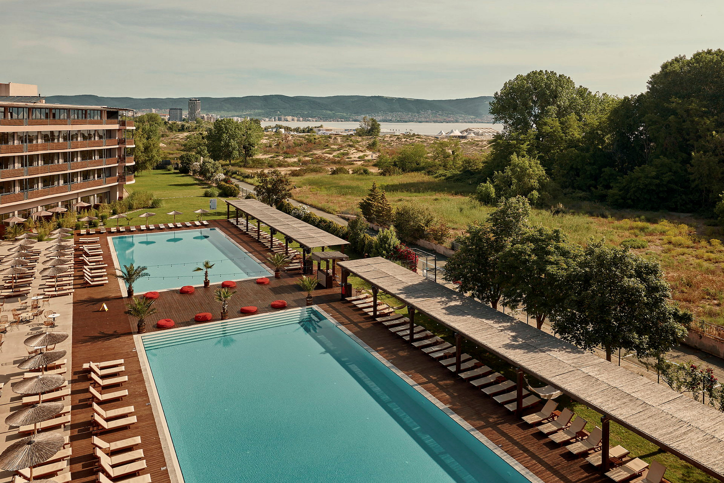 Stor pool med liggestole og parasoller ved siden af et hotel, med grønt område og landskab i baggrunden. (AI-genereret tekst)