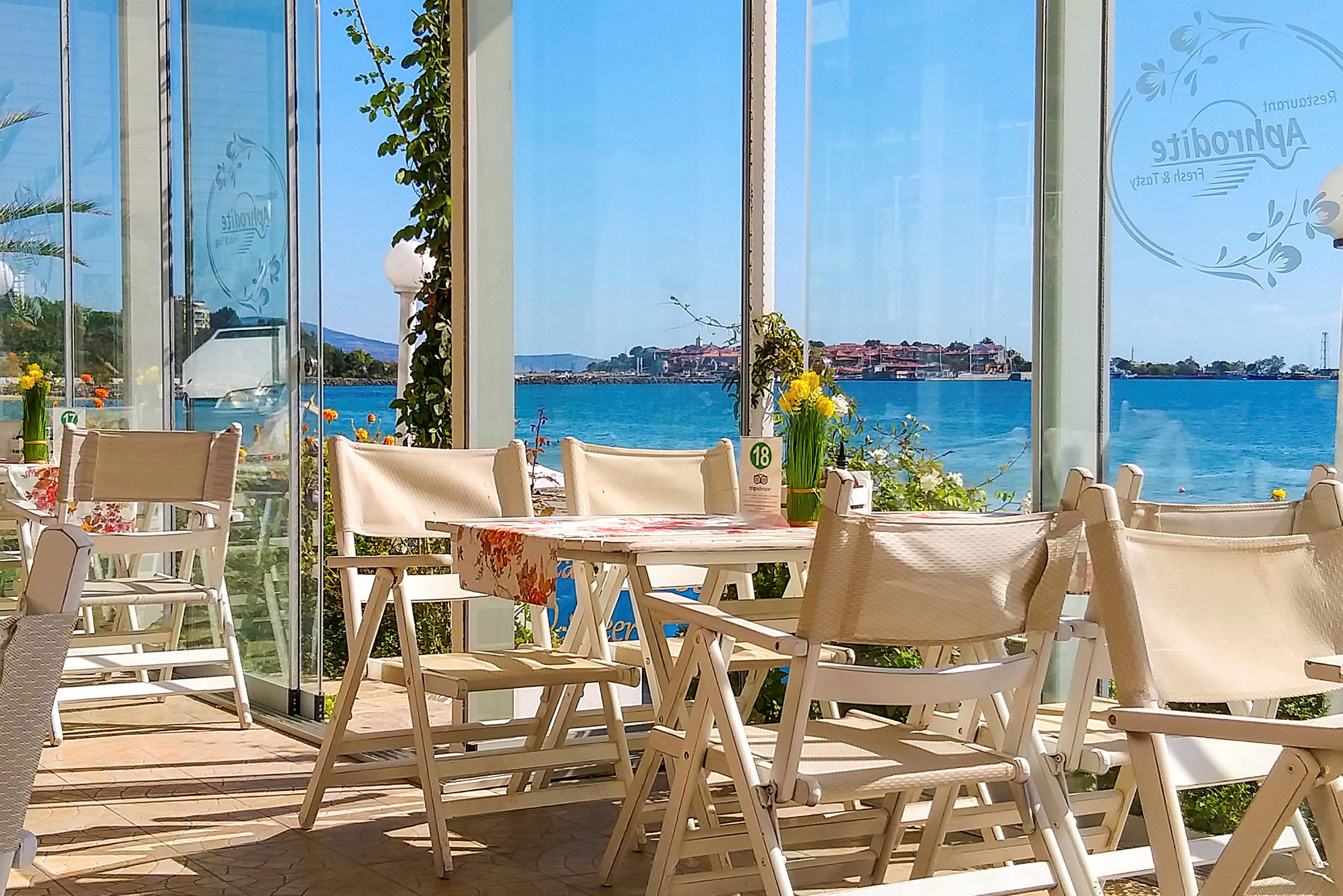 Udsigt fra en restaurant med hvide stole og borde, med glasvægge der giver udsigt over havet og en kystby. (AI-genereret tekst)