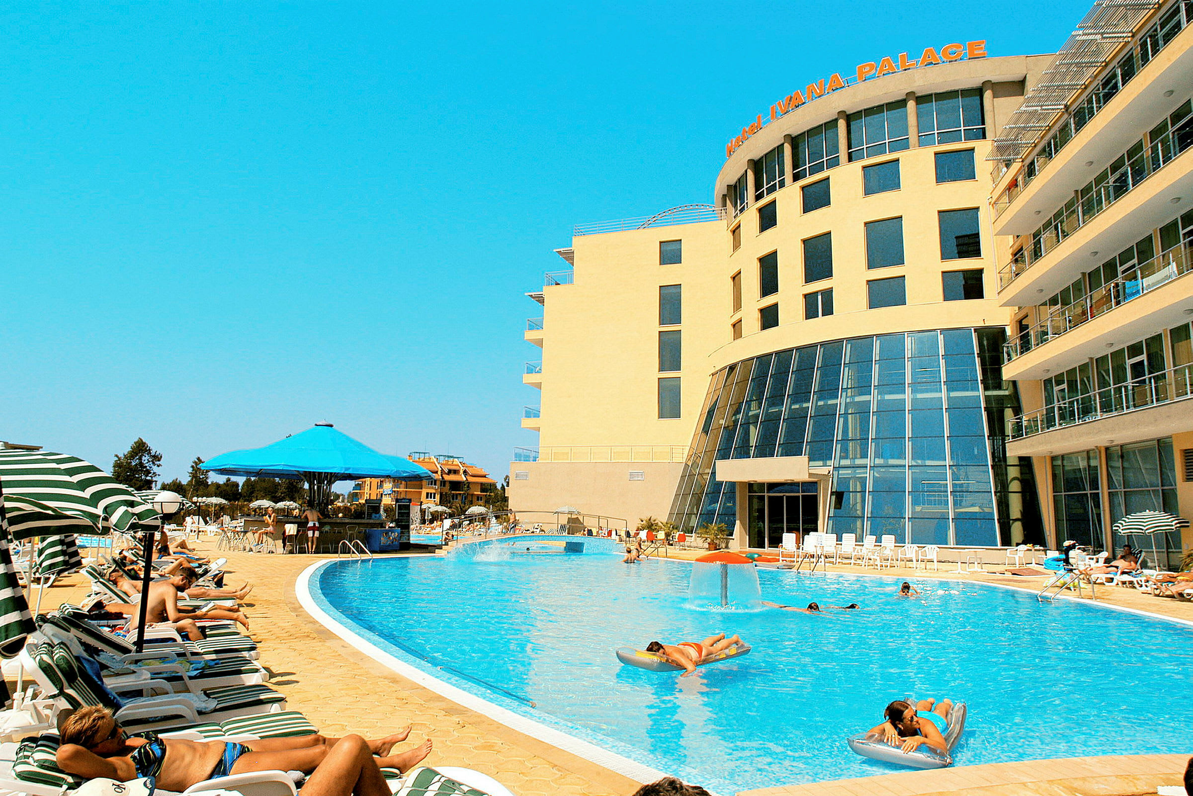 Et hotel med en stor udendørs pool og gæster, der slapper af på liggestole omkring den. (AI-genereret tekst)