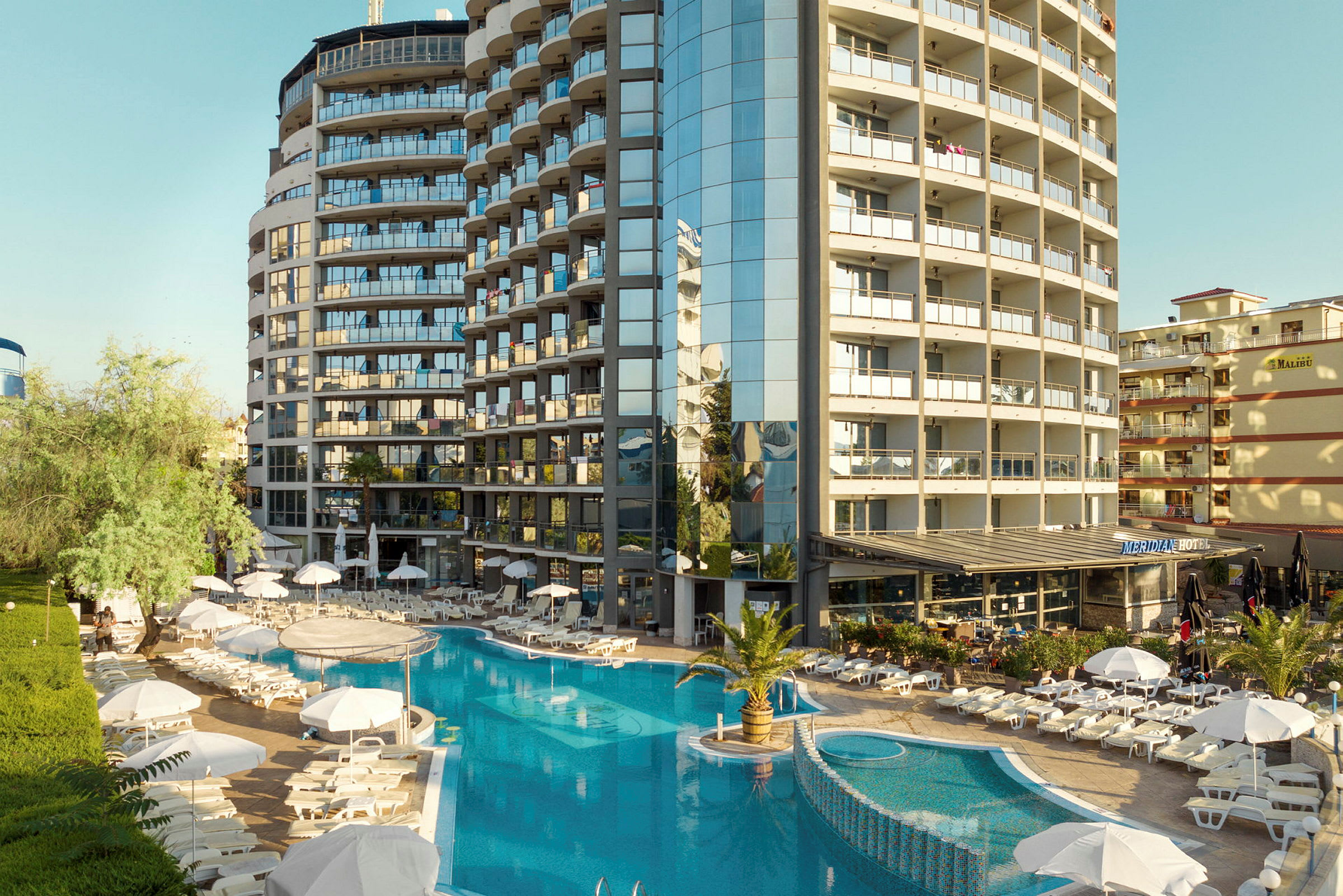 Et hotelkompleks med en pool omgivet af liggestole og parasoller. (AI-genereret tekst)