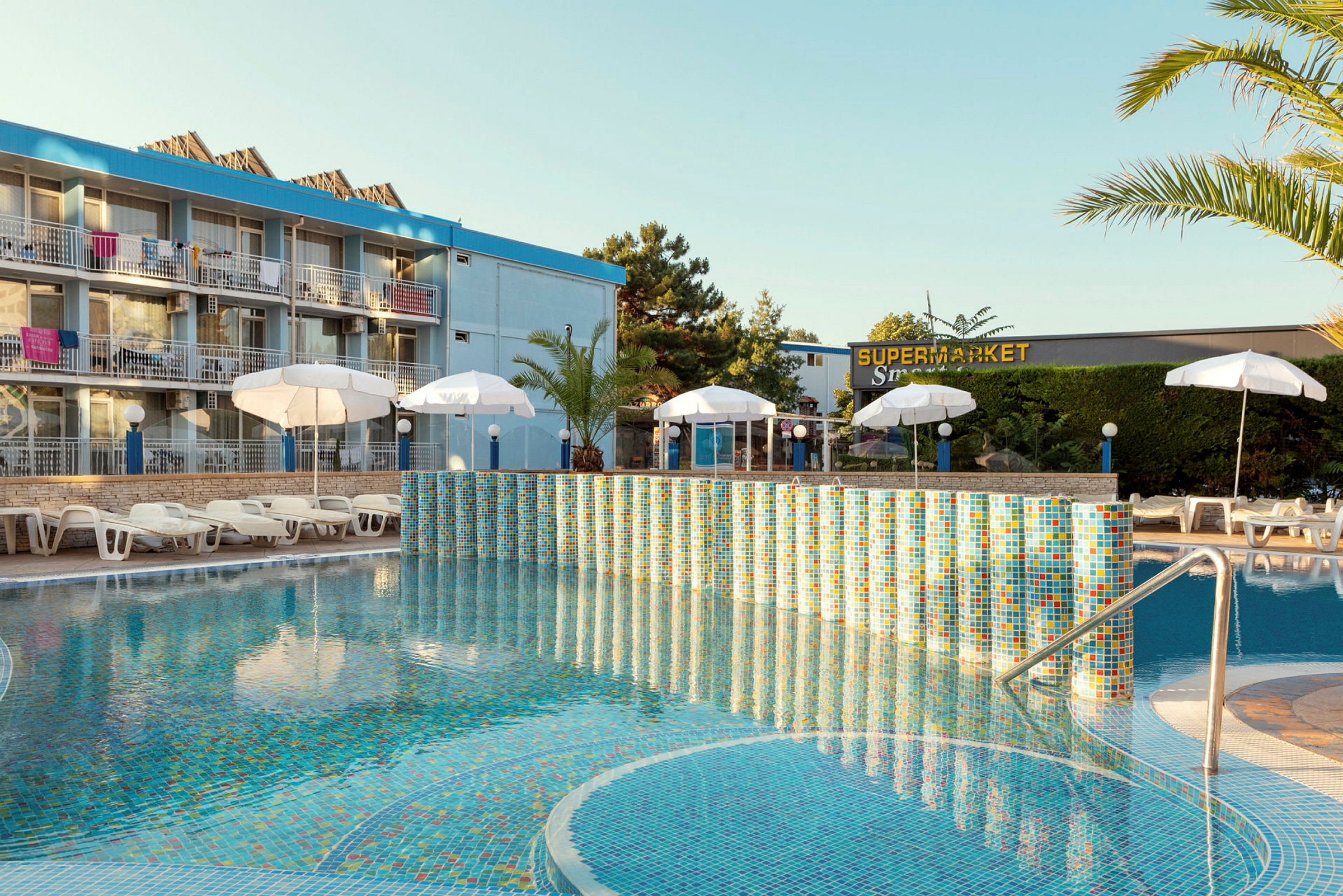 Hotel med swimmingpool, liggestole og parasoller foran bygningen. (AI-genereret tekst)