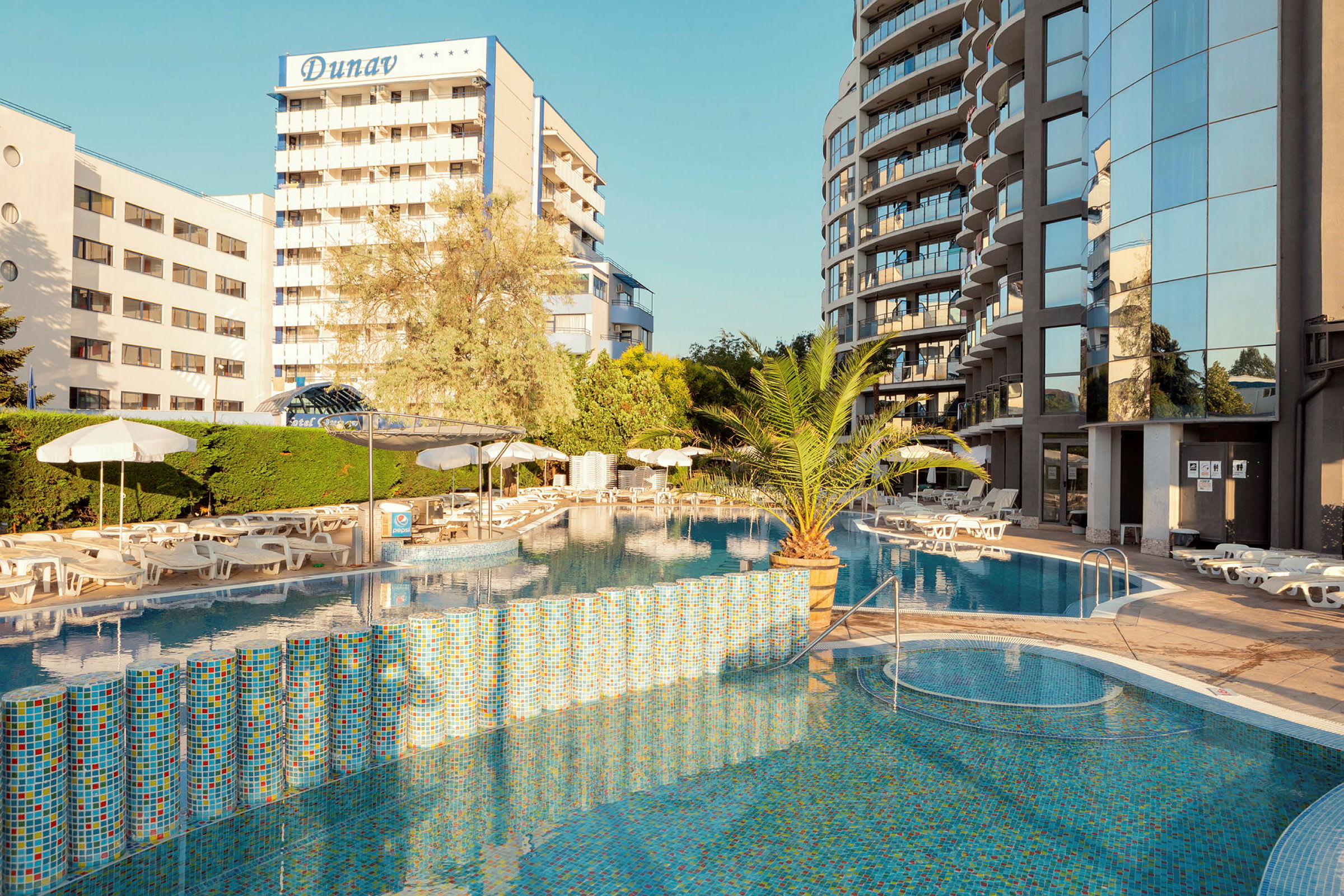 Billede af hotelområde med udendørs pool, omgivet af liggestole og parasoller. Baggrund med bygninger og beplantning. (AI-genereret tekst)