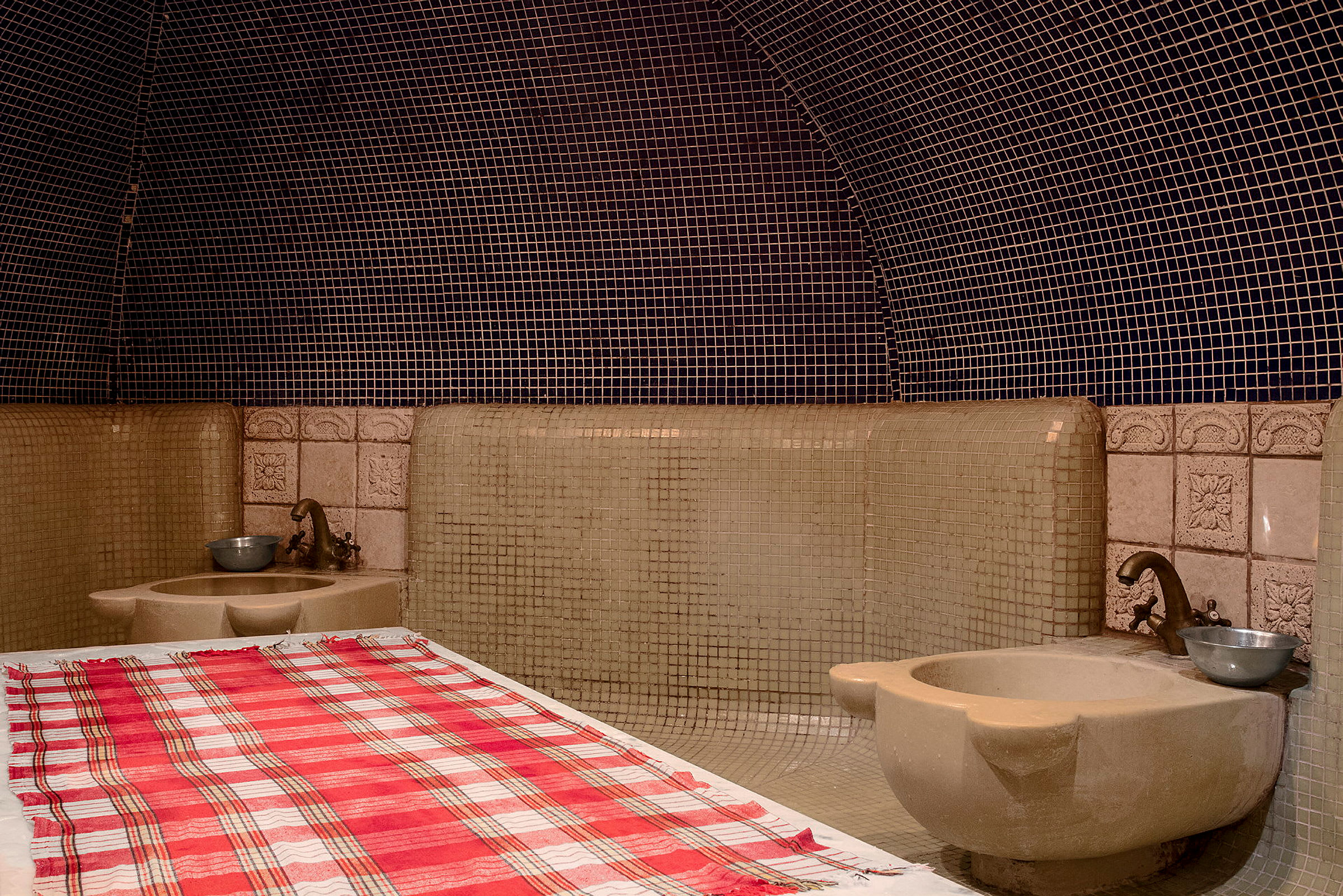 Et traditionelt hamam-rum med flisebeklædte vægge og en bænk dækket af et ternet håndklæde. (AI-genereret tekst)