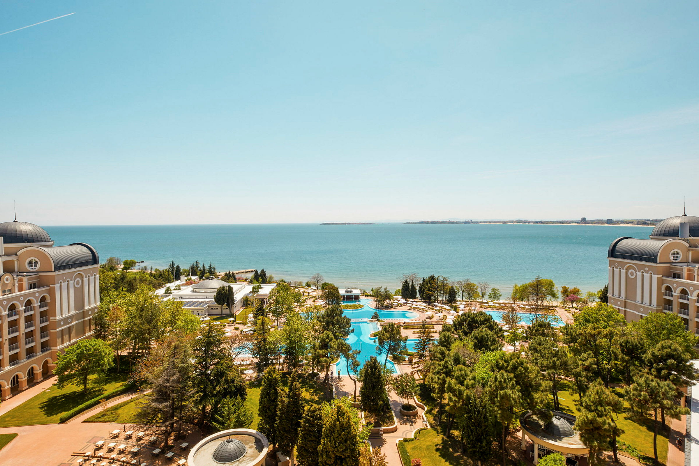 Udsigt over et hotelområde med to bygninger, swimmingpools, grønne områder og havet i baggrunden. (AI-genereret tekst)