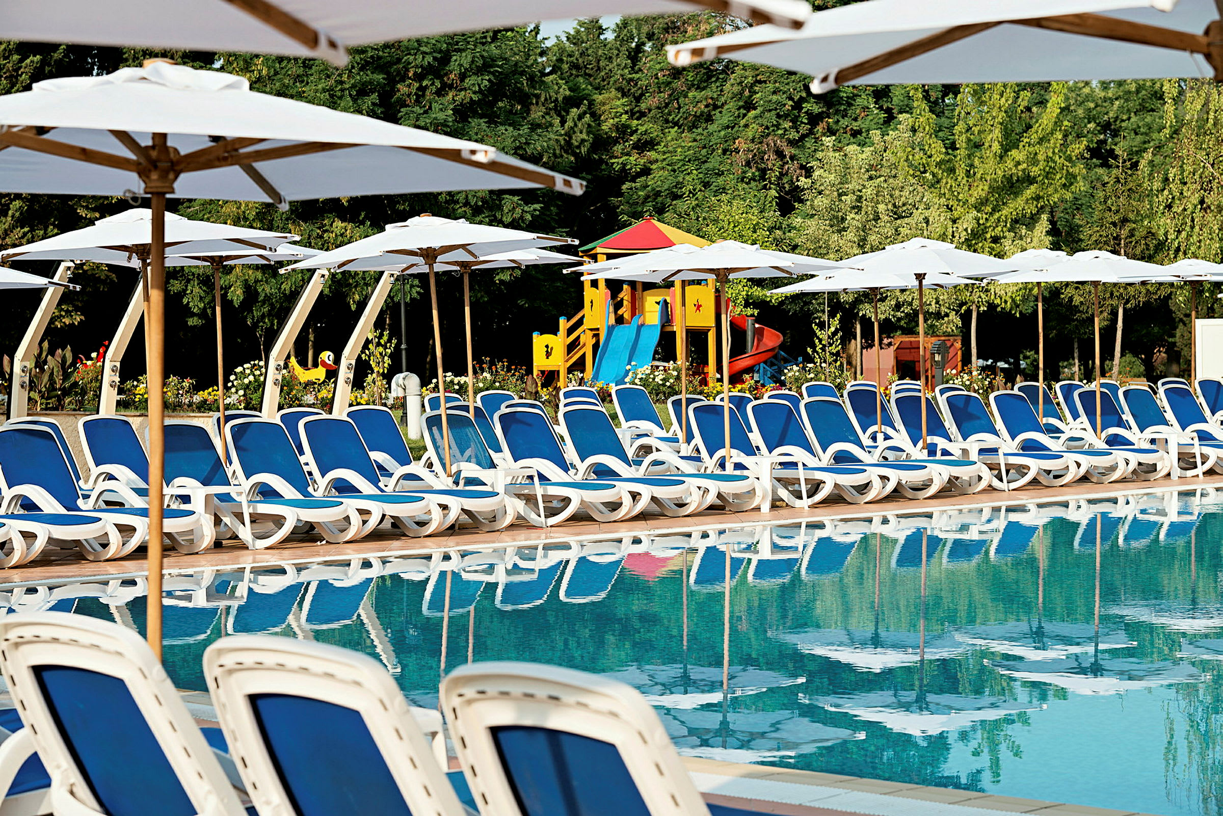 Billede af liggestole med blå hynder ved en swimmingpool. Bag liggestolene ses hvide parasoller og en legeplads for børn. (AI-genereret tekst)