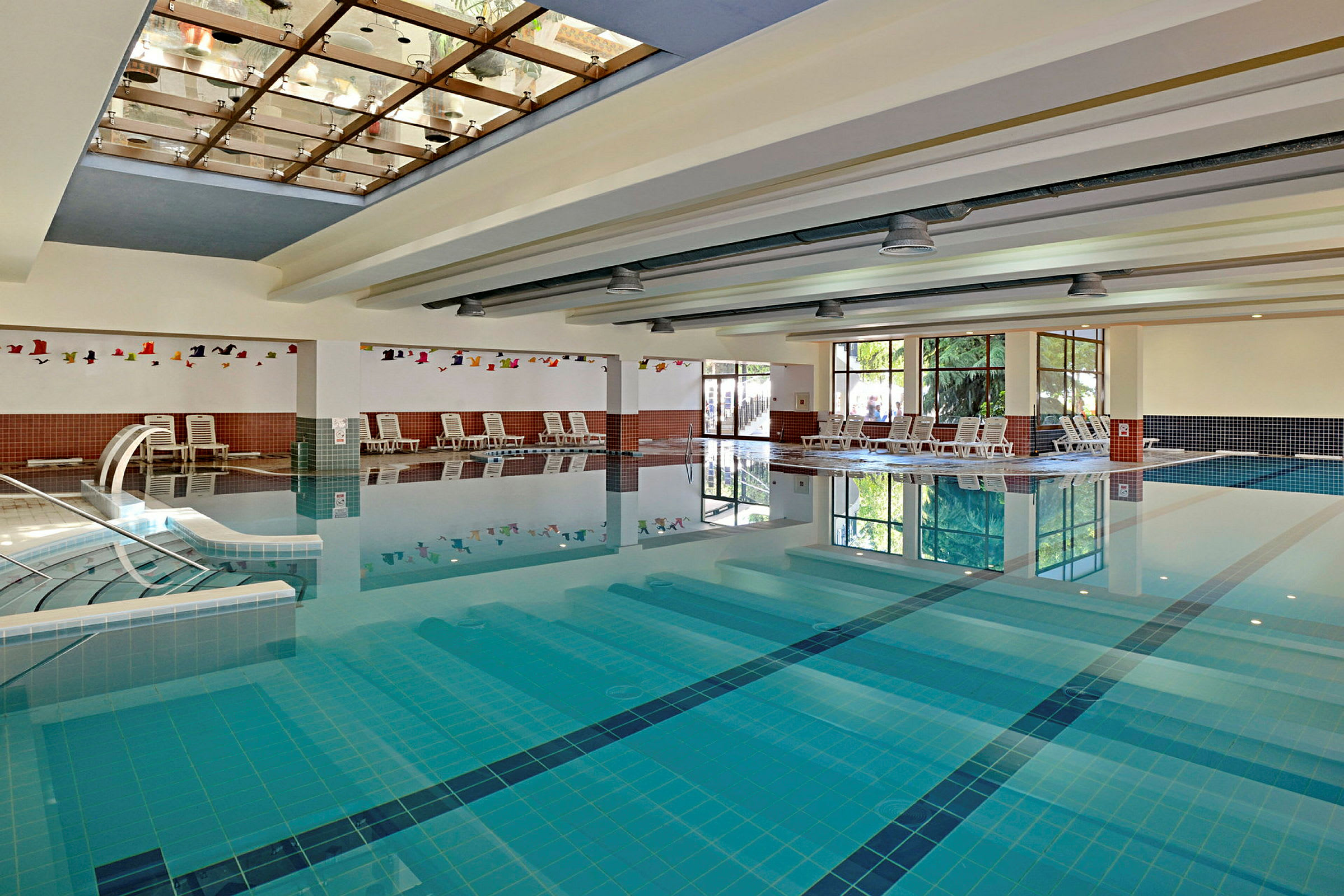 Indendørs swimmingpool med liggestole langs kanten og glaspartier, der lader naturligt lys komme ind. (AI-genereret tekst)