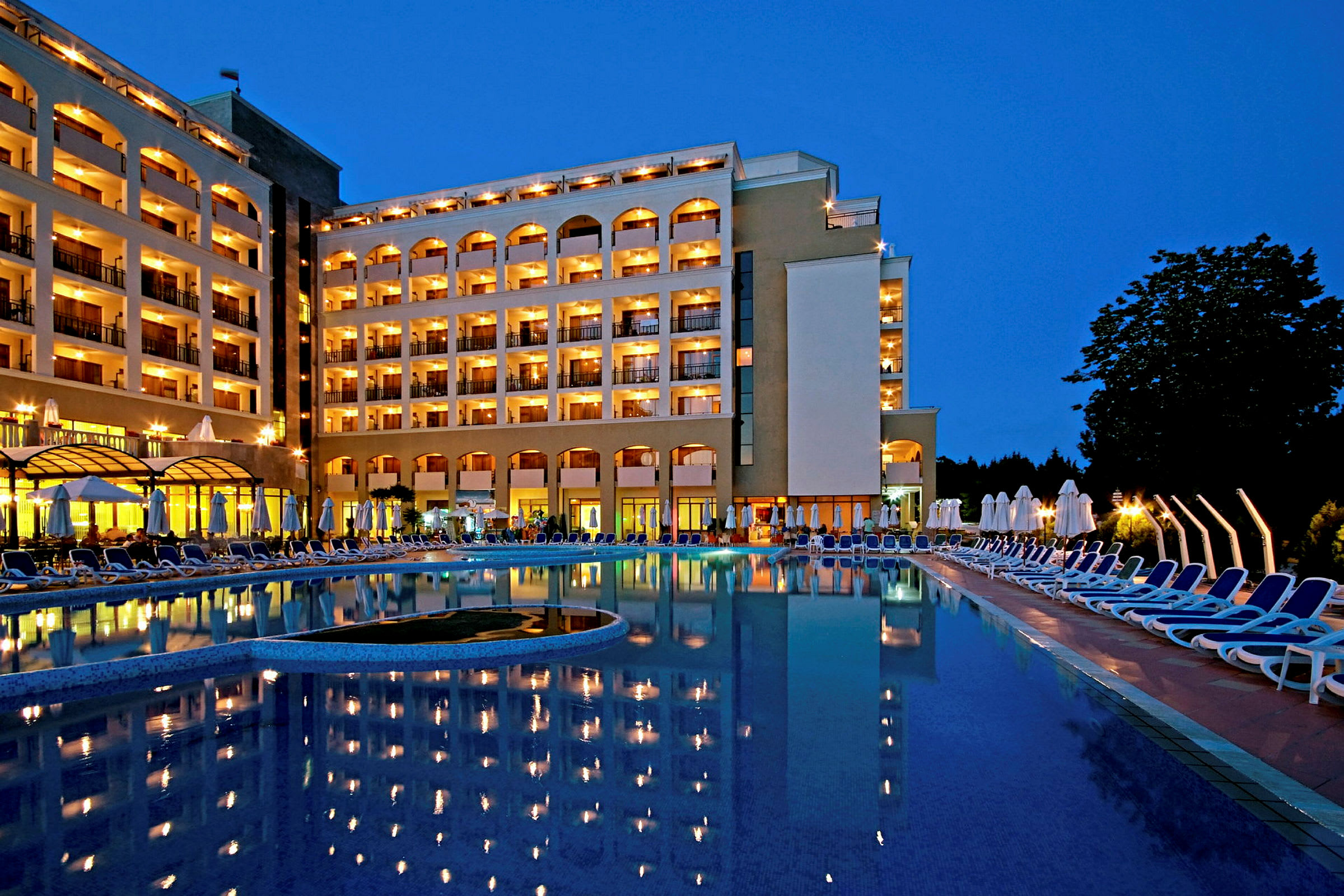 Et aftenbillede af en oplyst hotelbygning ved en pool, der reflekterer lysene. (AI-genereret tekst)