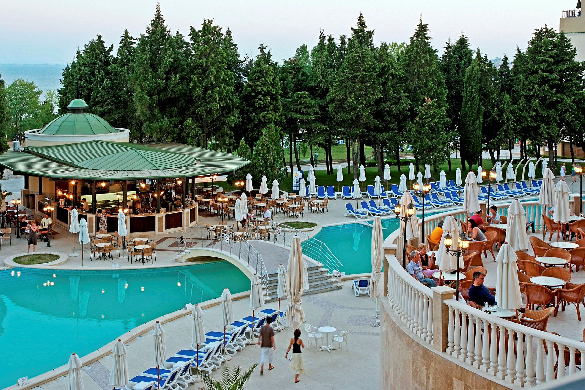 Billede af et hotelområde med en pool, omgivet af liggestole og en udendørs café med siddepladser. Træer og grønne områder i baggrunden. (AI-genereret tekst)