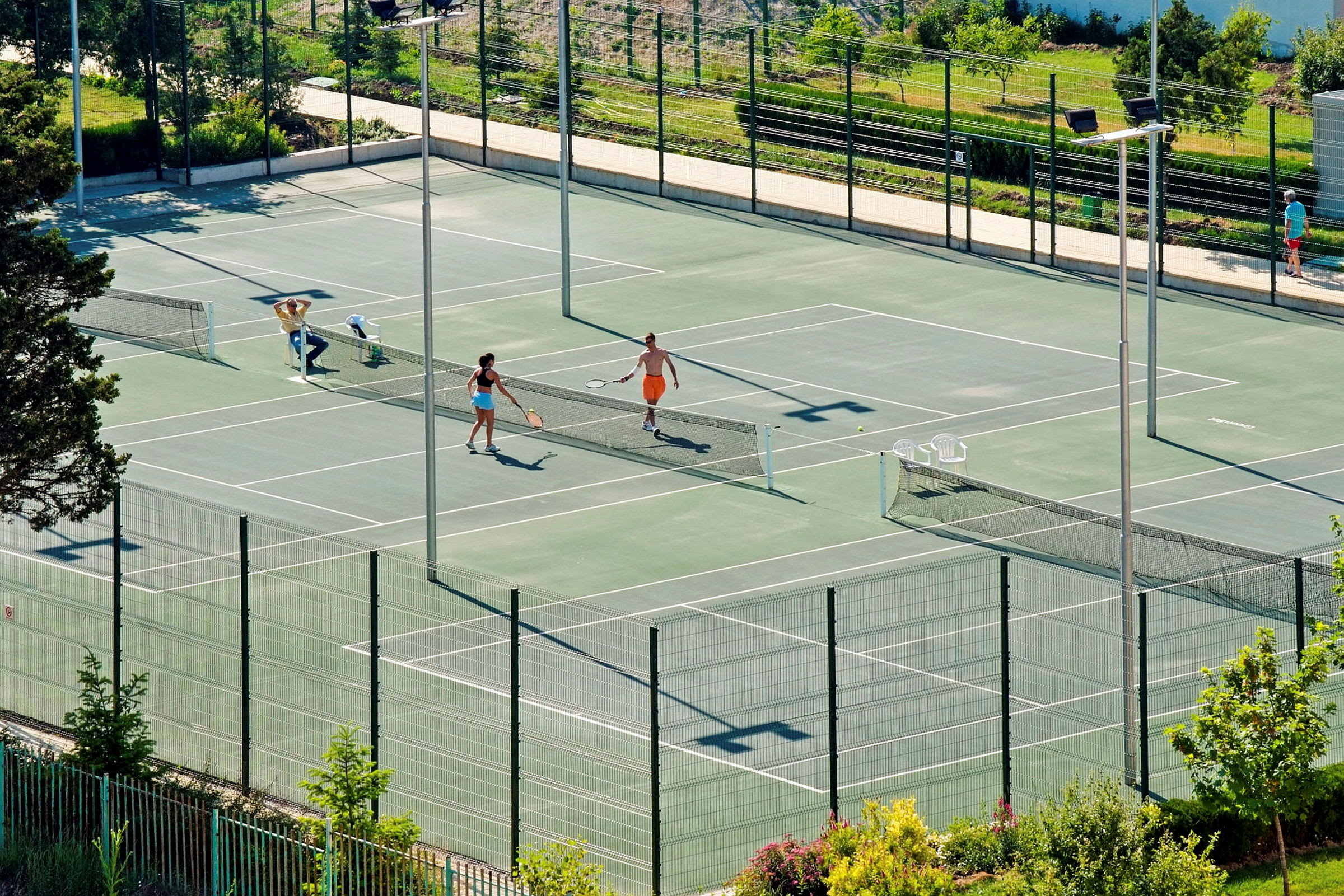 To personer spiller tennis på en udendørs bane omgivet af grønne omgivelser. (AI-genereret tekst)