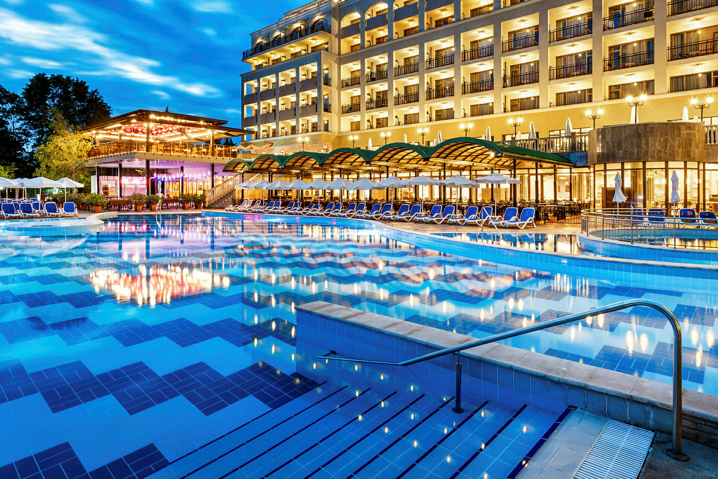 Et hotel med mange altaner og områder til afslapning ved en stor oplyst udendørs pool. (AI-genereret tekst)