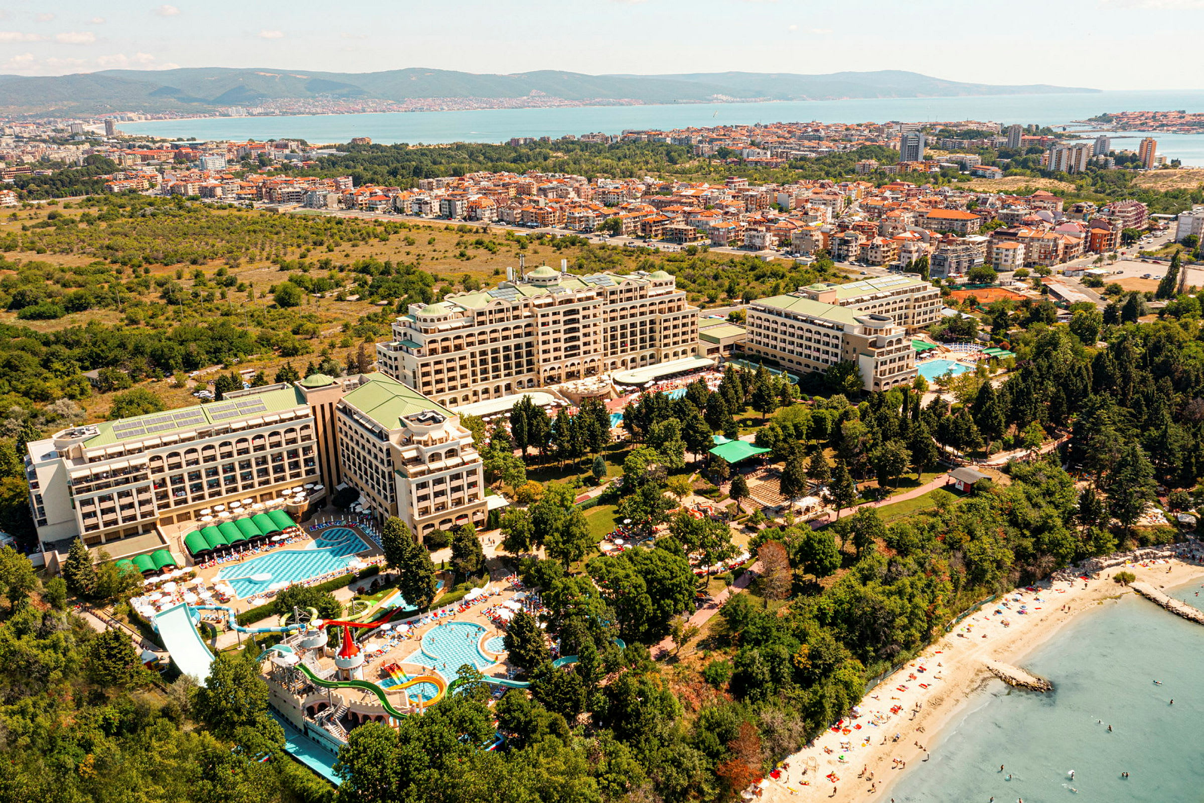 Udsigt over et feriested med flere hotelbygninger, swimmingpools, grønne områder og en strand ved havet. (AI-genereret tekst)