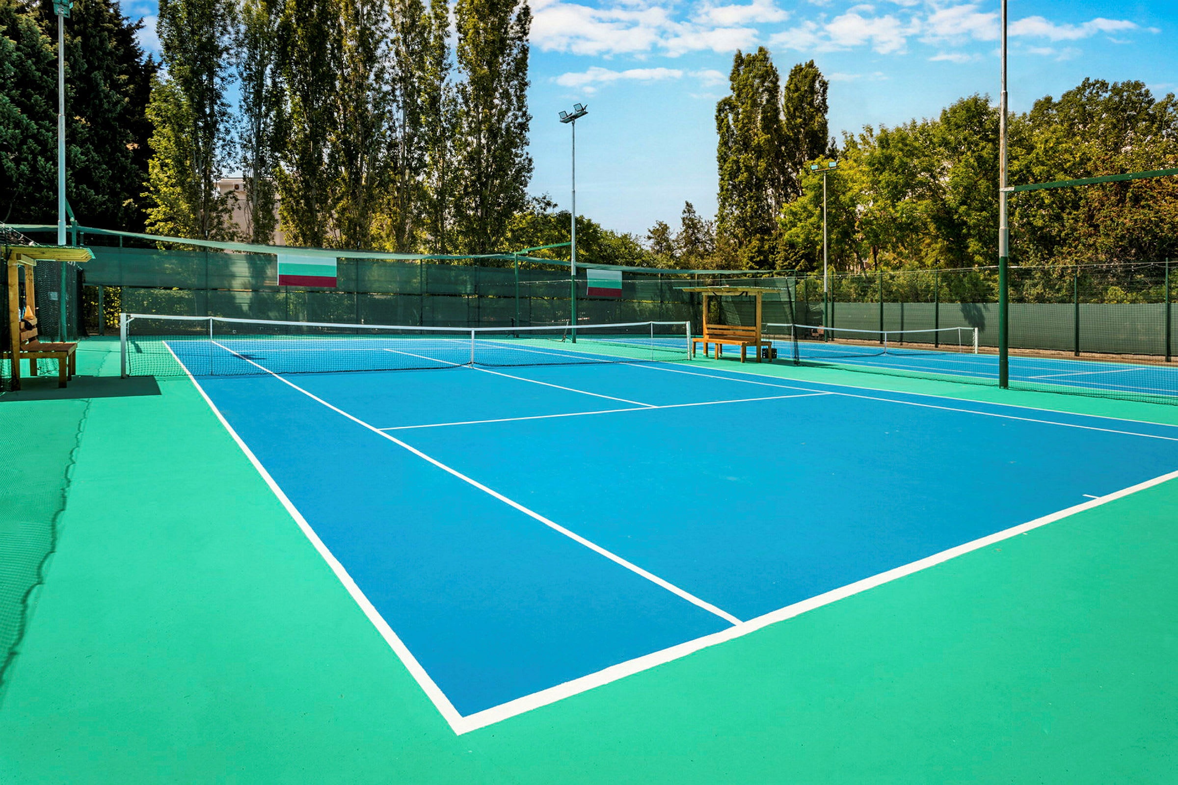 Billede af en tennisbane med blå overflade og hvide linjer, omgivet af træer og hegn. (AI-genereret tekst)