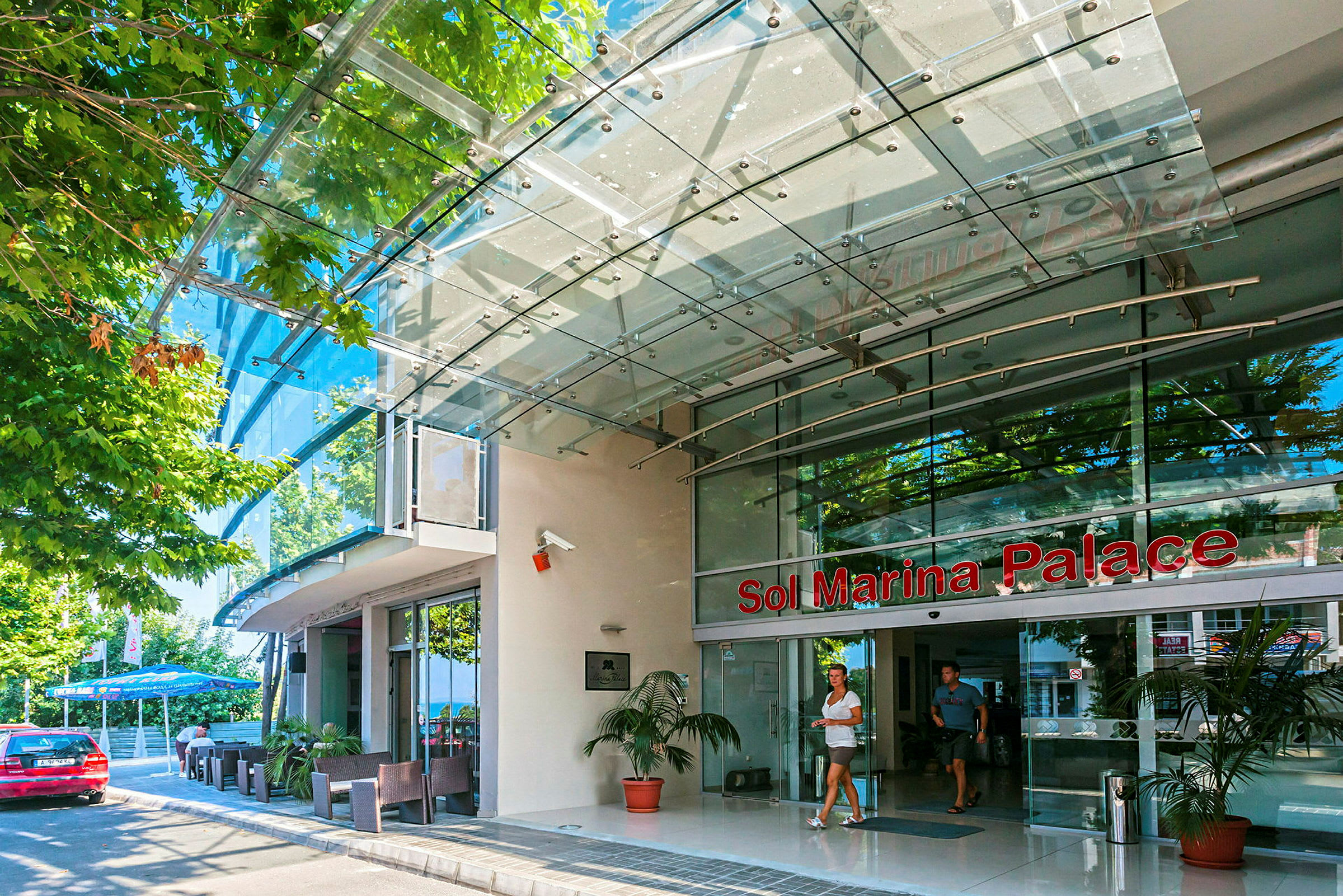 Indgangen til Sol Marina Palace med glasfacade og et skilt med navnet. (AI-genereret tekst)