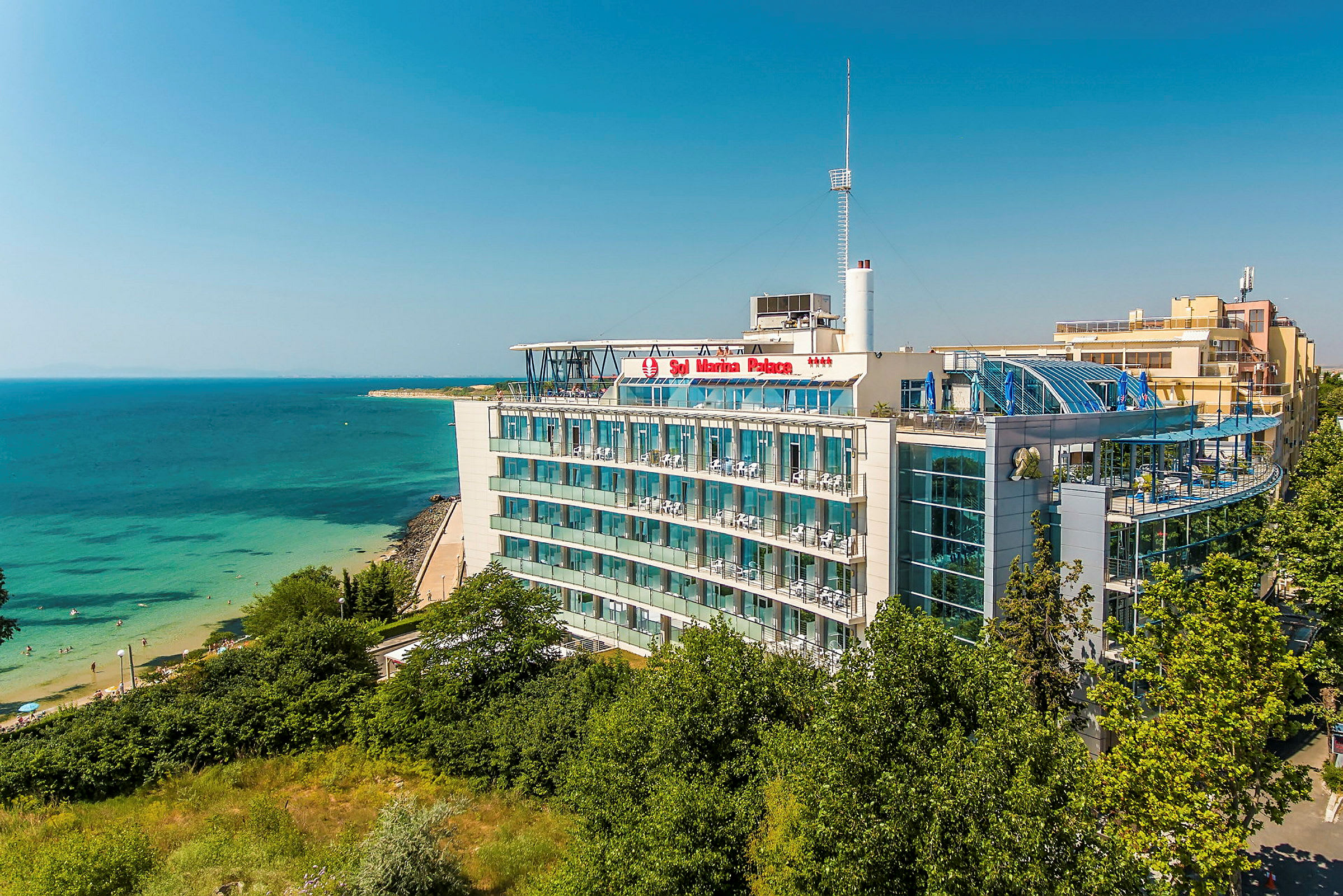 Et hotel ved havet med altaner og glasfacader, omgivet af grønne områder, med en strand i nærheden. (AI-genereret tekst)