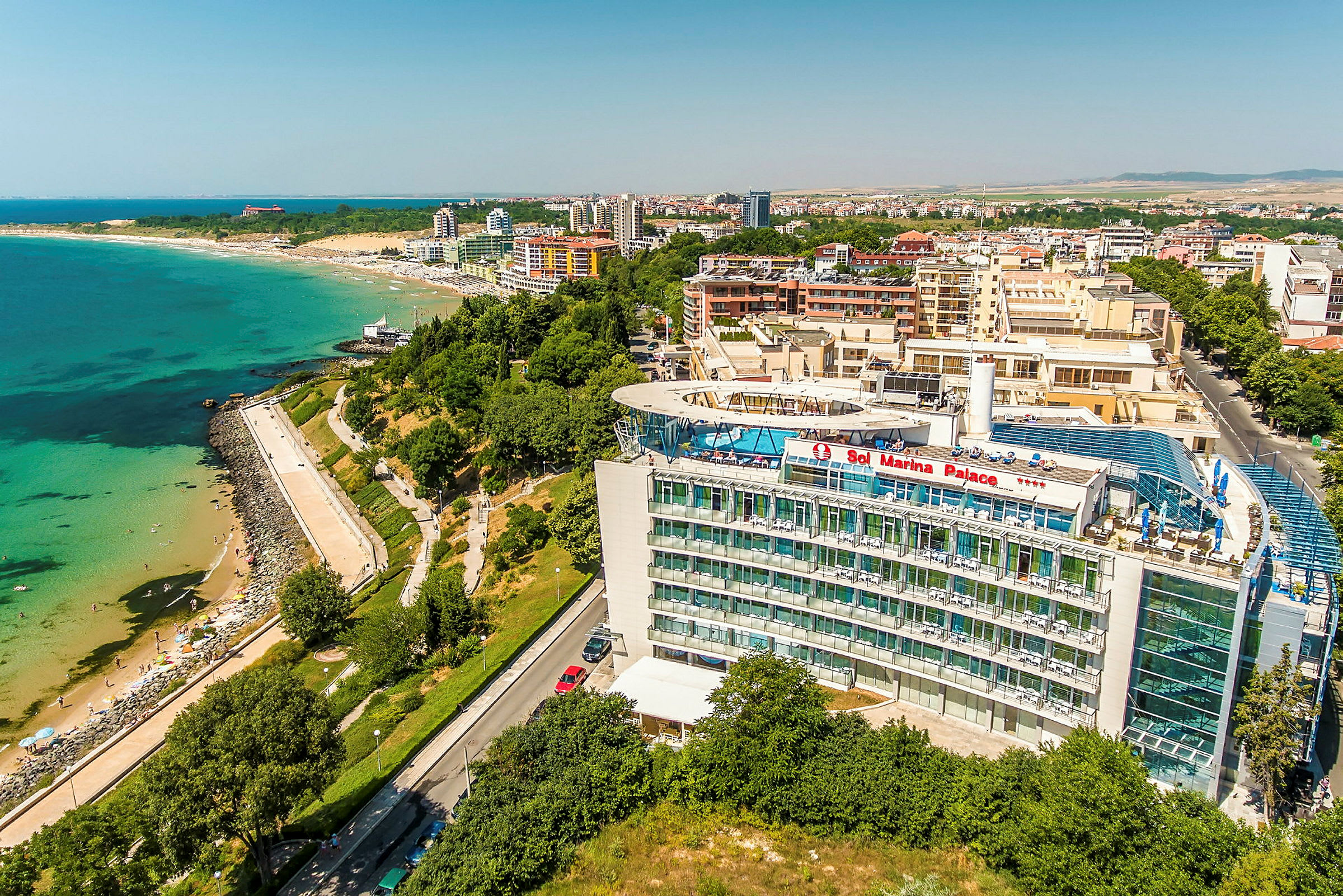 Luftfoto af et hotel med teksten "Sol Marina Palace", beliggende nær en strand og kystlinje med beplantning og bygninger i baggrunden. (AI-genereret tekst)