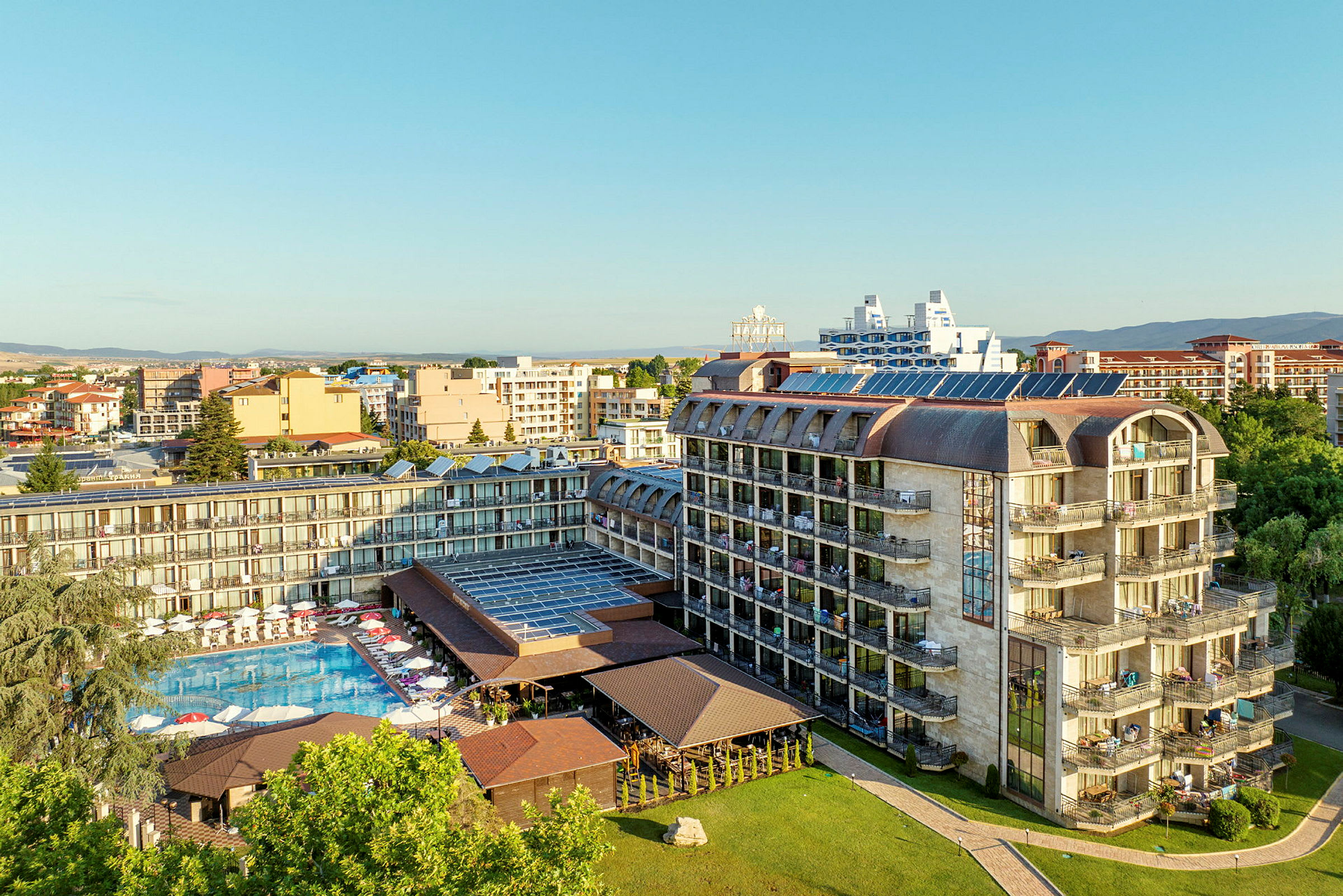 Luftfoto af et hotel med swimmingpool og liggestole, omgivet af andre bygninger og grønne områder. (AI-genereret tekst)