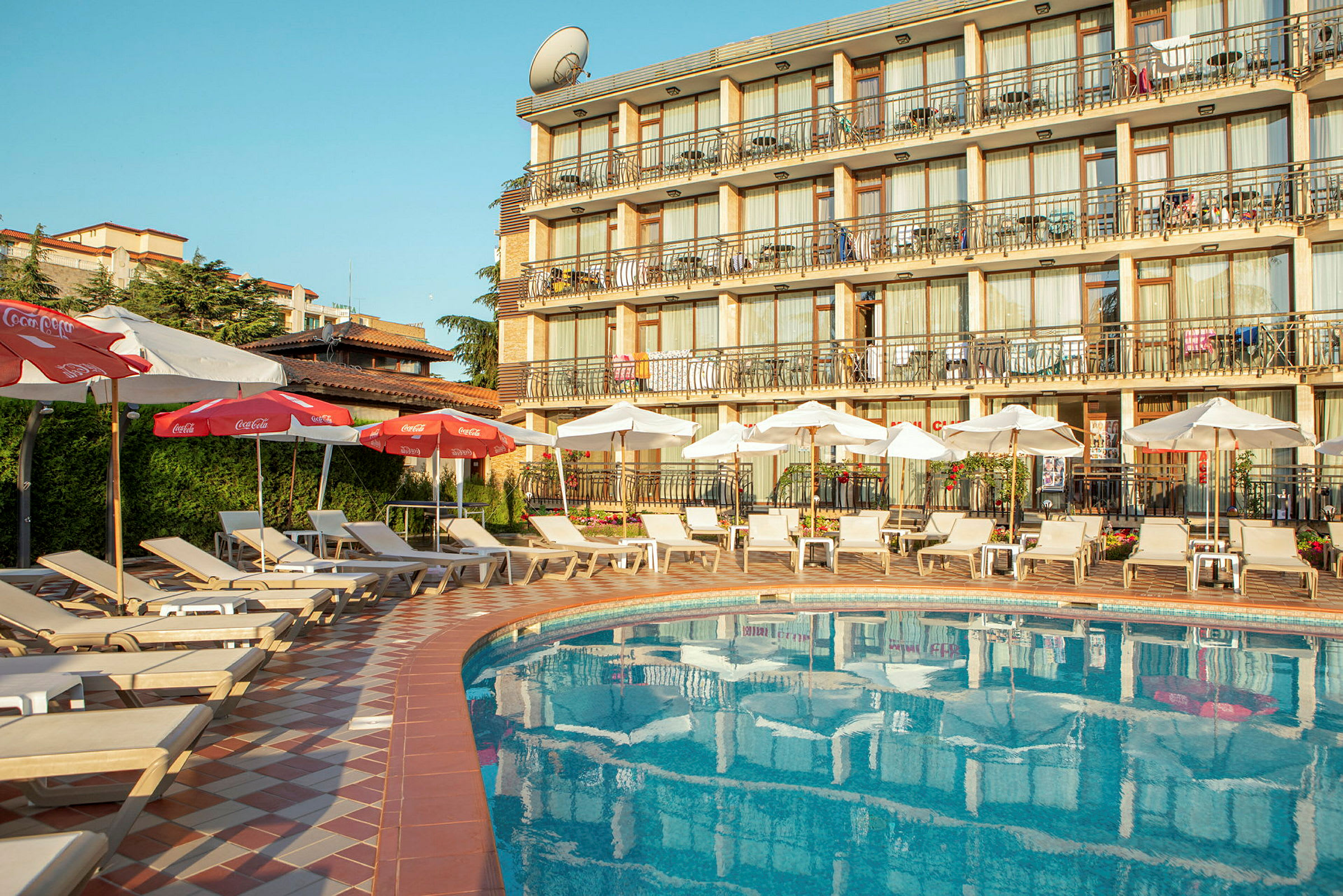Billede af en hotelpool med liggestole og parasoller samt hotelbygning i baggrunden. (AI-genereret tekst)