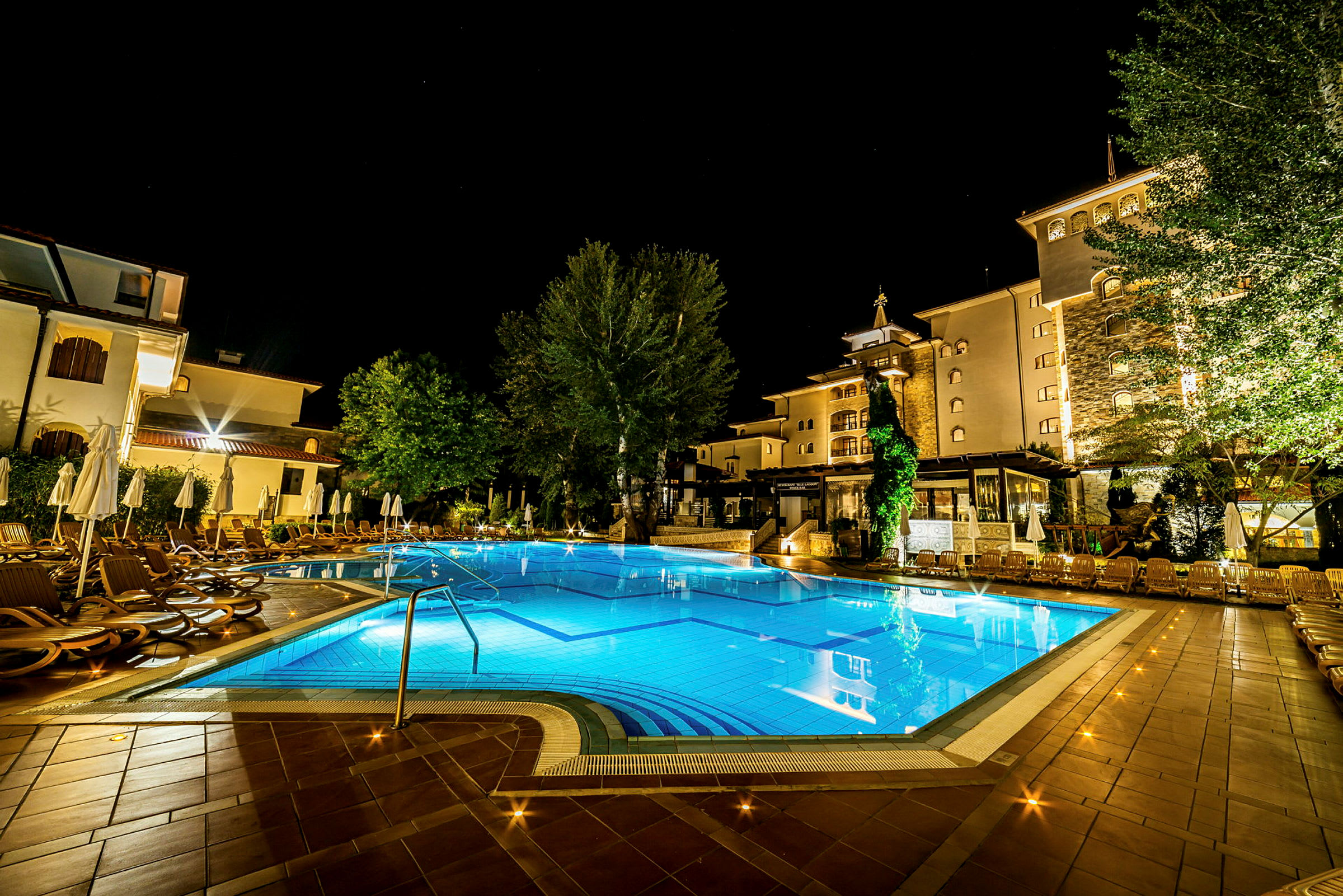 Et natbillede af en oplyst hotelpool omgivet af liggestole og parasoller, med hotelbygninger i baggrunden. (AI-genereret tekst)