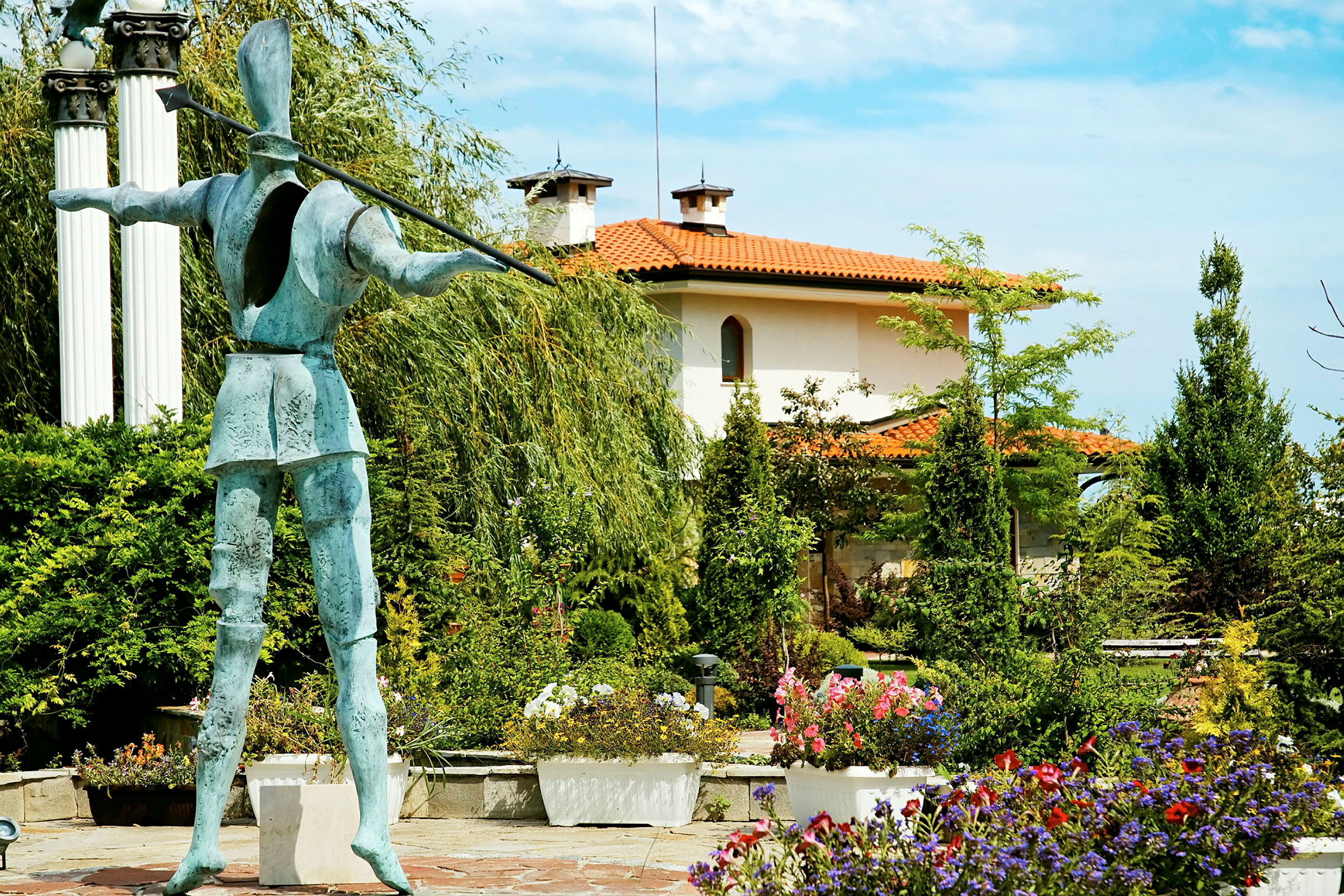 En statue i en have omgivet af blomster og grønne buske. I baggrunden ses et hus med orange tag. (AI-genereret tekst)