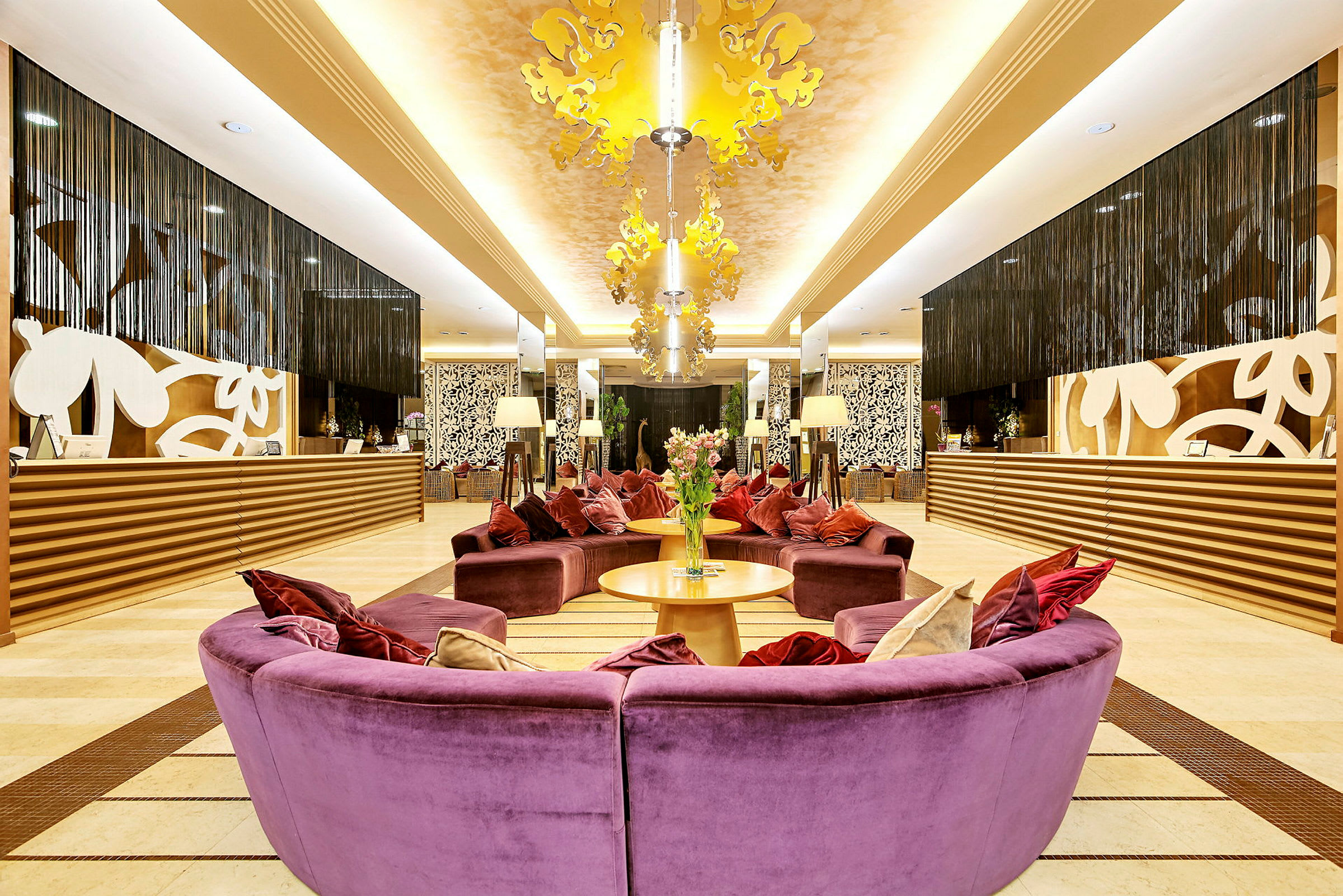 Receptionen på et hotel med elegant interiør. Farverige sofaer i lilla med puder er centreret sammen med to borde og blomster i midten. Bag receptionen ses dekorative mønstre og hængende gardiner. (AI-genereret tekst)