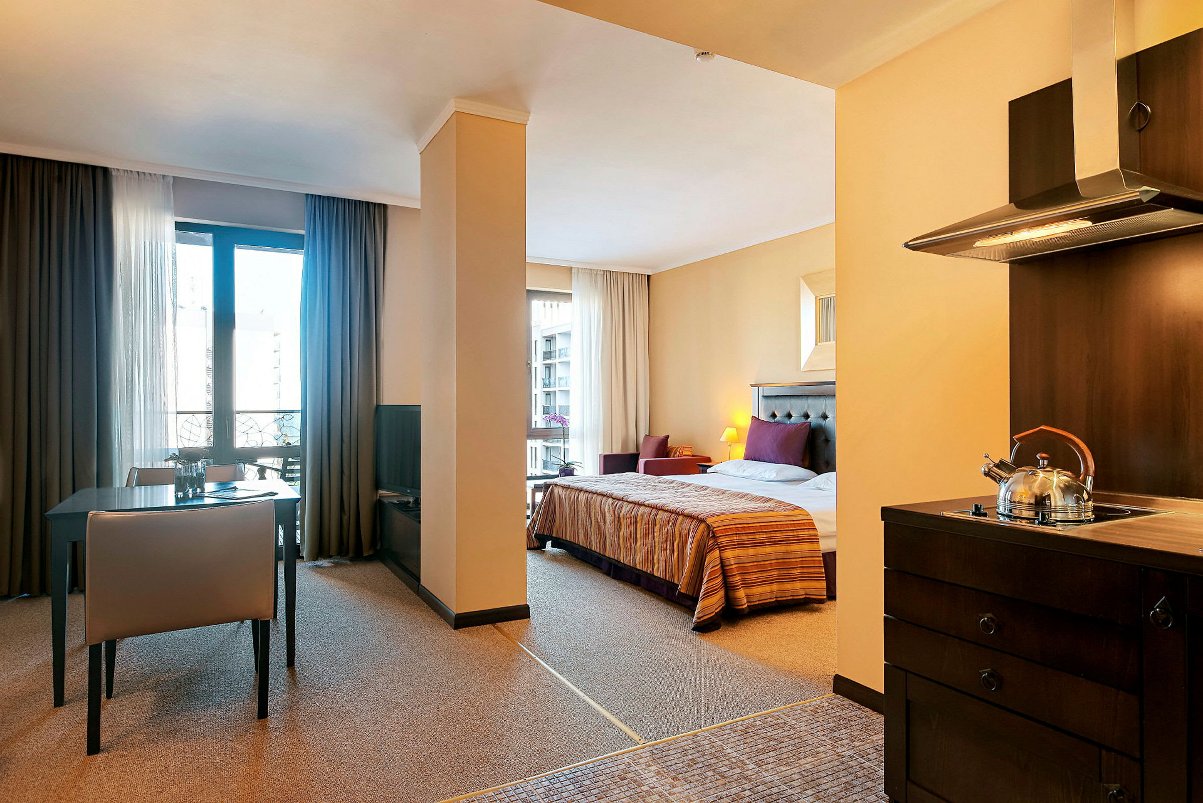 Interiør af et hotelværelse med soveområde, spisebord og køkken. Sengen er redt, og der er en tekande på komfuret. (AI-genereret tekst)