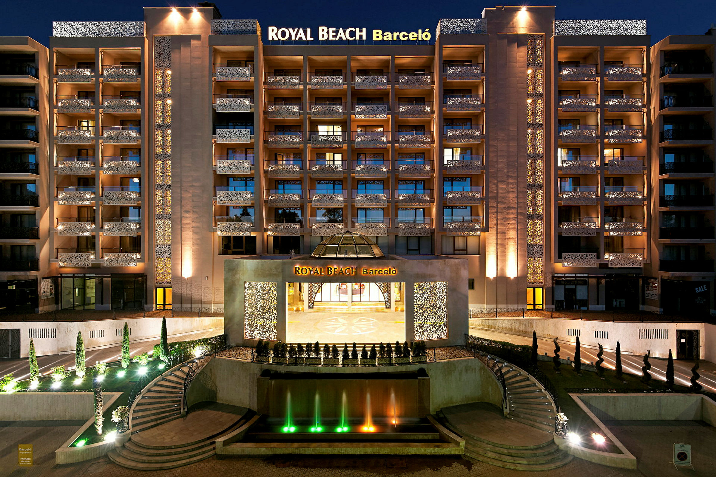 Et natbillede af et oplyst hotel med navnet Royal Beach Barceló, med en springvand og trapper i forgrunden. (AI-genereret tekst)