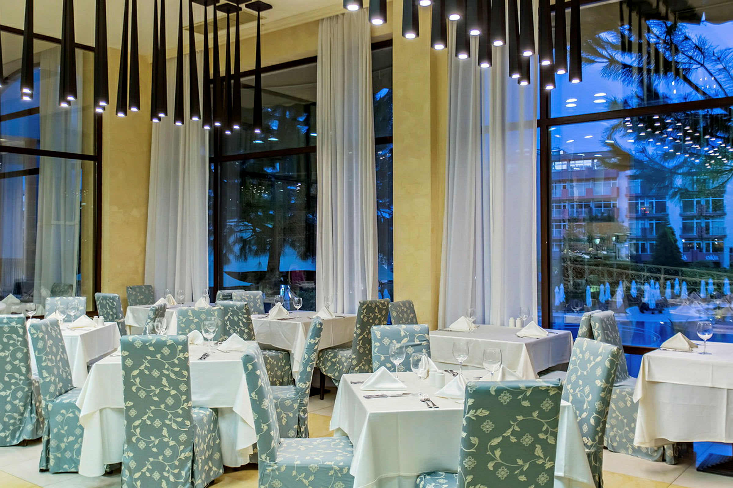 En elegant restaurant med borde dækket med hvide duge og stole med blomstrede mønstre, store vinduer med hvide gardiner. (AI-genereret tekst)