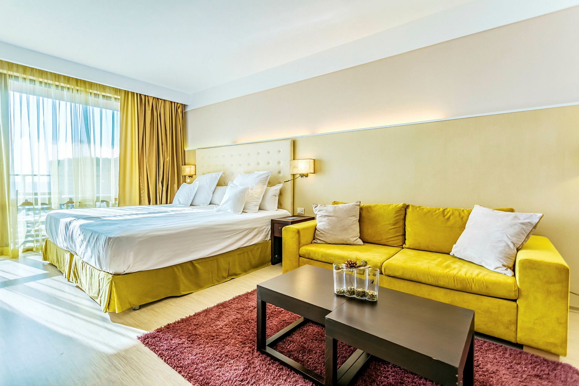 Et hotelværelse med en stor dobbeltseng, gul sofa, et bord med dekorativ vase og et rødt tæppe. (AI-genereret tekst)