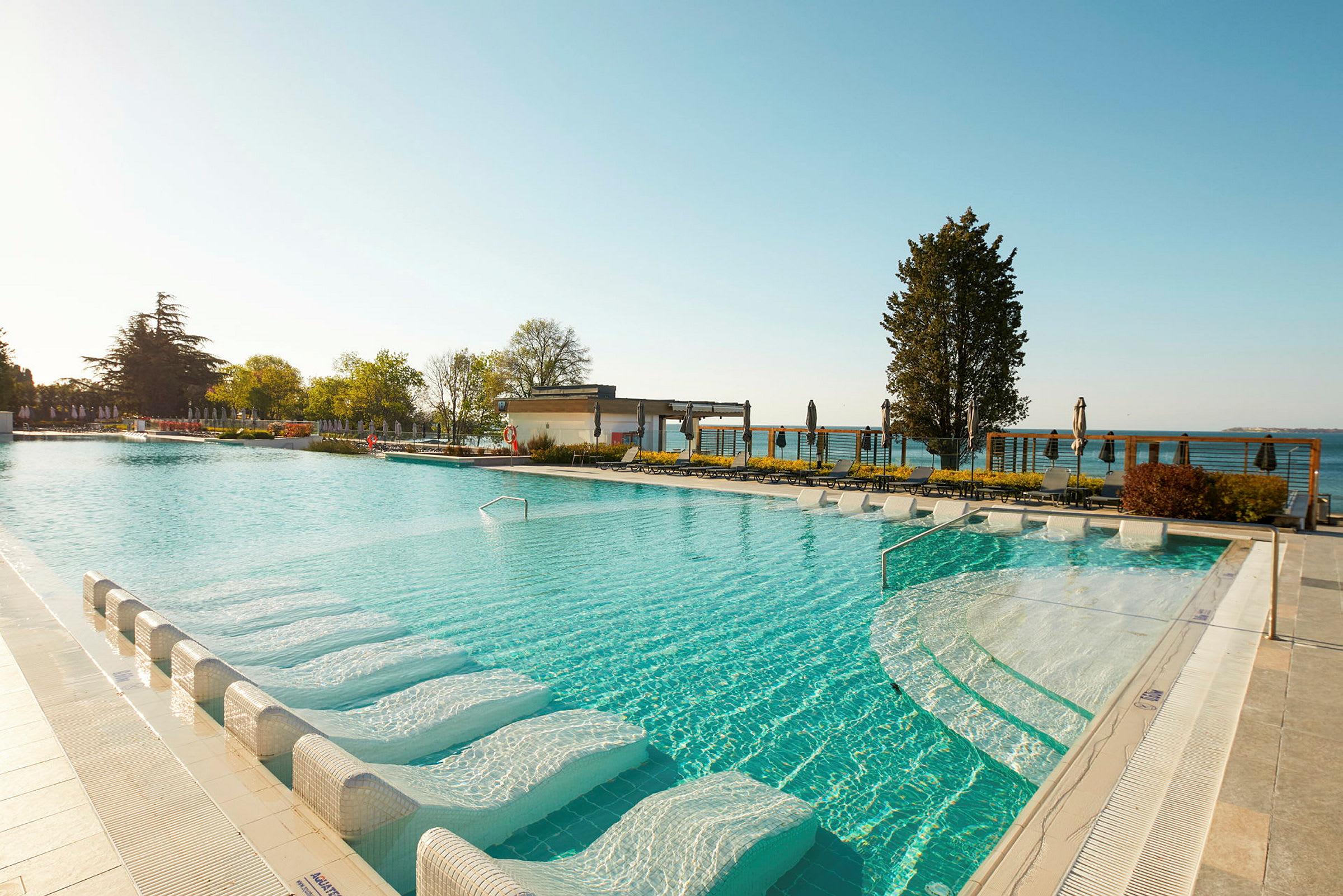 Moderne swimmingpool med liggestole i vandet og udsigt over de omkringliggende grønne områder og himmel. (AI-genereret tekst)