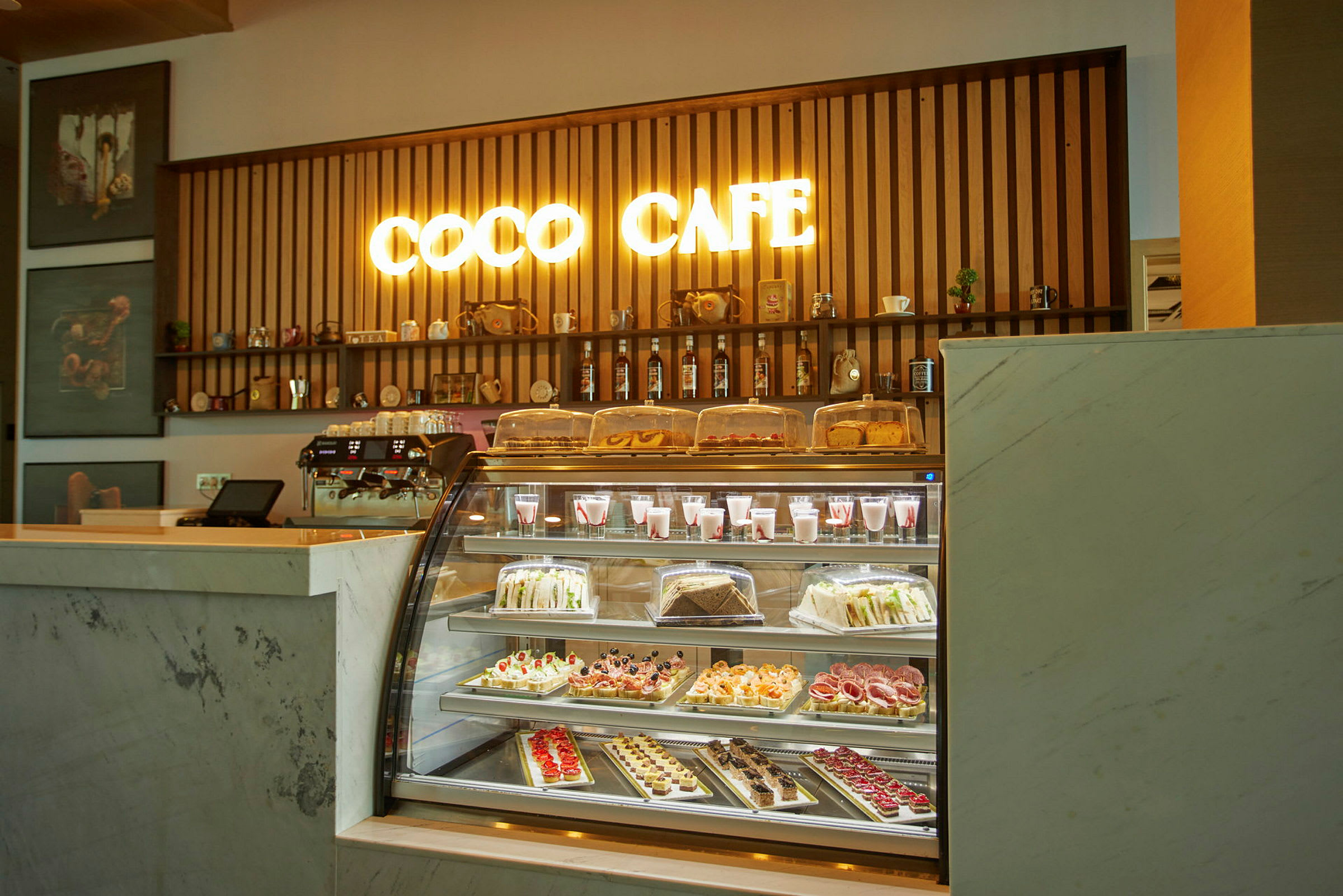 En cafédisk med et oplyst skilt med teksten 'Coco Cafe' og et udvalg af bagværk og drikkevarer bag glas. (AI-genereret tekst)