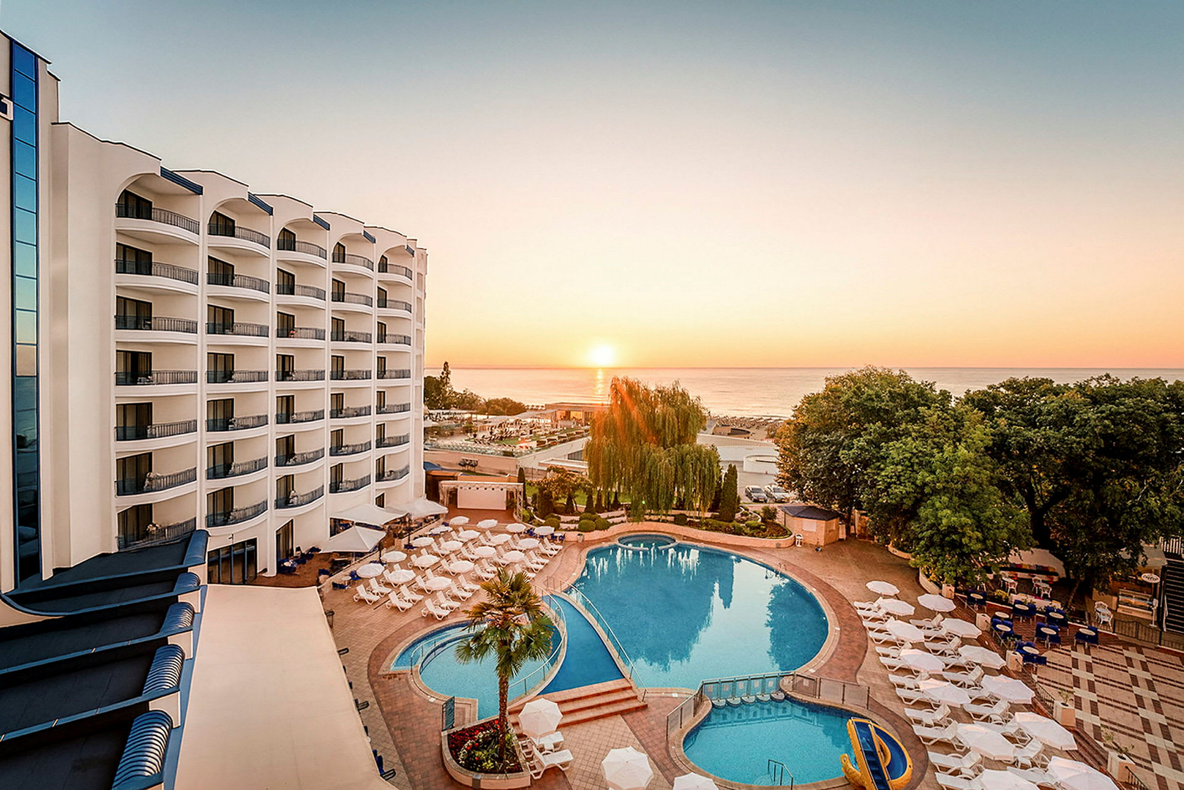 Billede af et hotelområde ved solnedgang med en stor pool omgivet af parasoller og liggestole samt havet i baggrunden. (AI-genereret tekst)