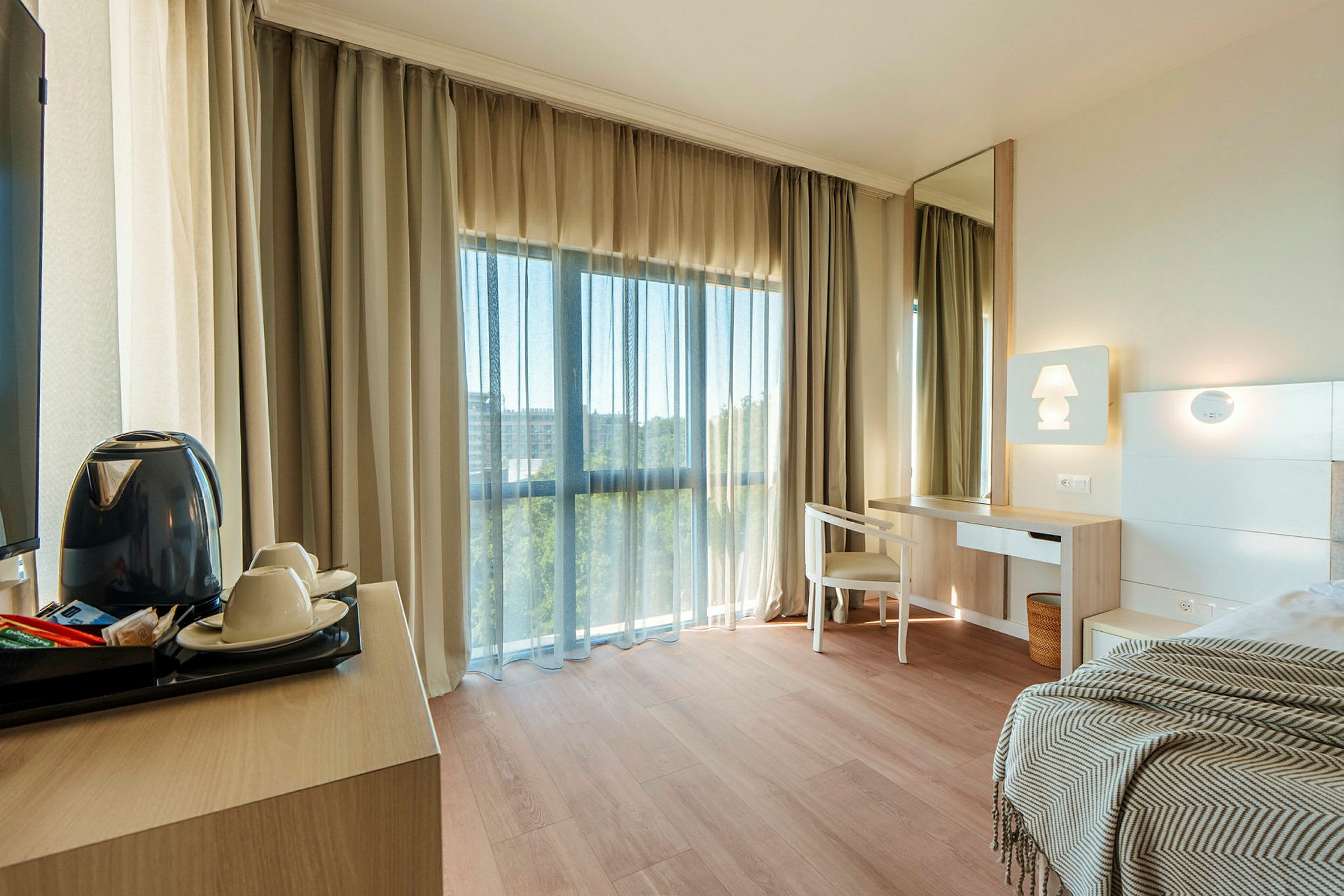 Et lyst hotelværelse med trægulv, et skrivebord med stol og spejl, samt en seng med et tæppe. Et stort vindue med gardiner lader dagslys komme ind. (AI-genereret tekst)
