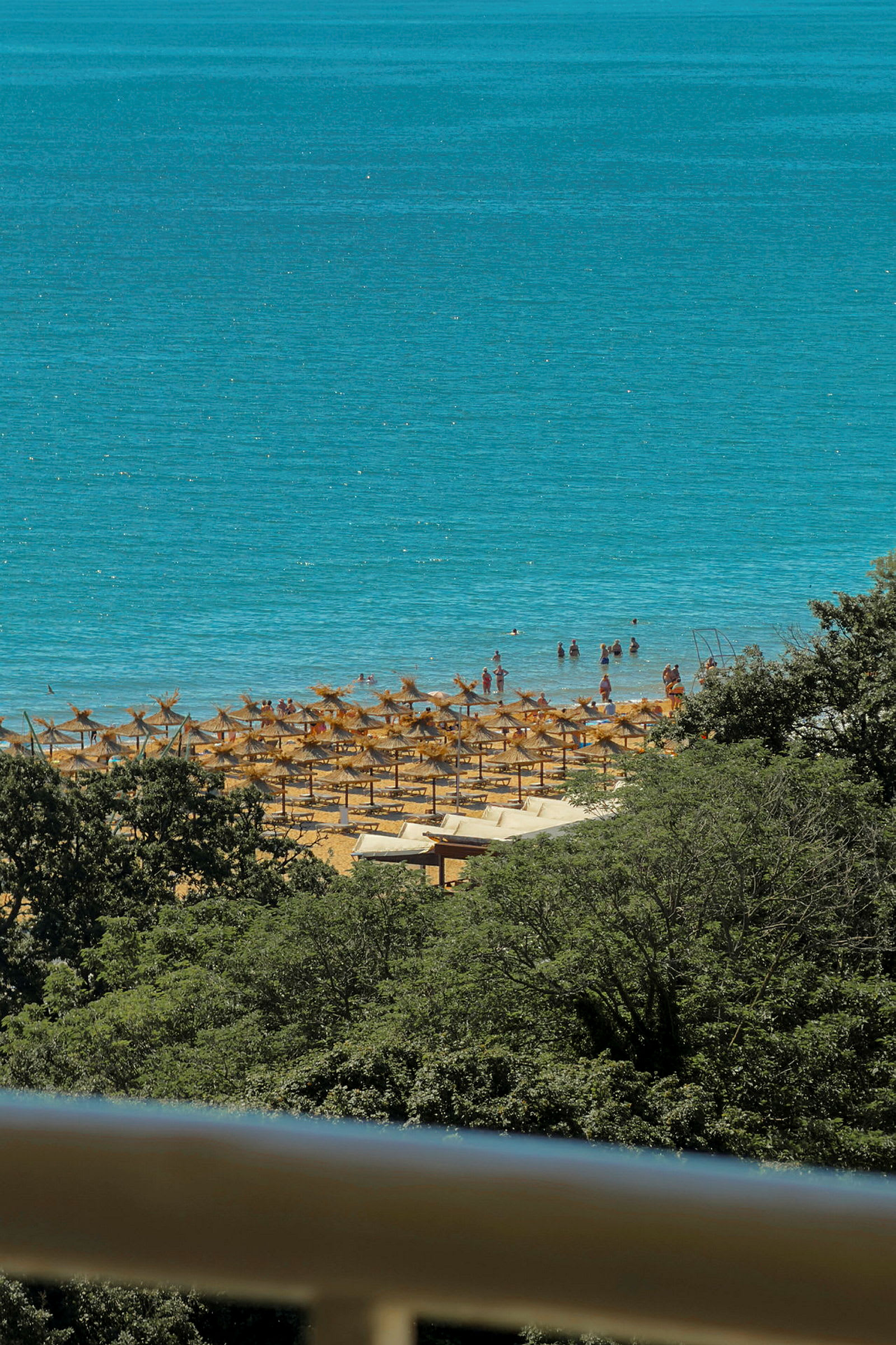 Udsigt over en strand med parasoller og mennesker, der bader, omgivet af grønne områder. (AI-genereret tekst)