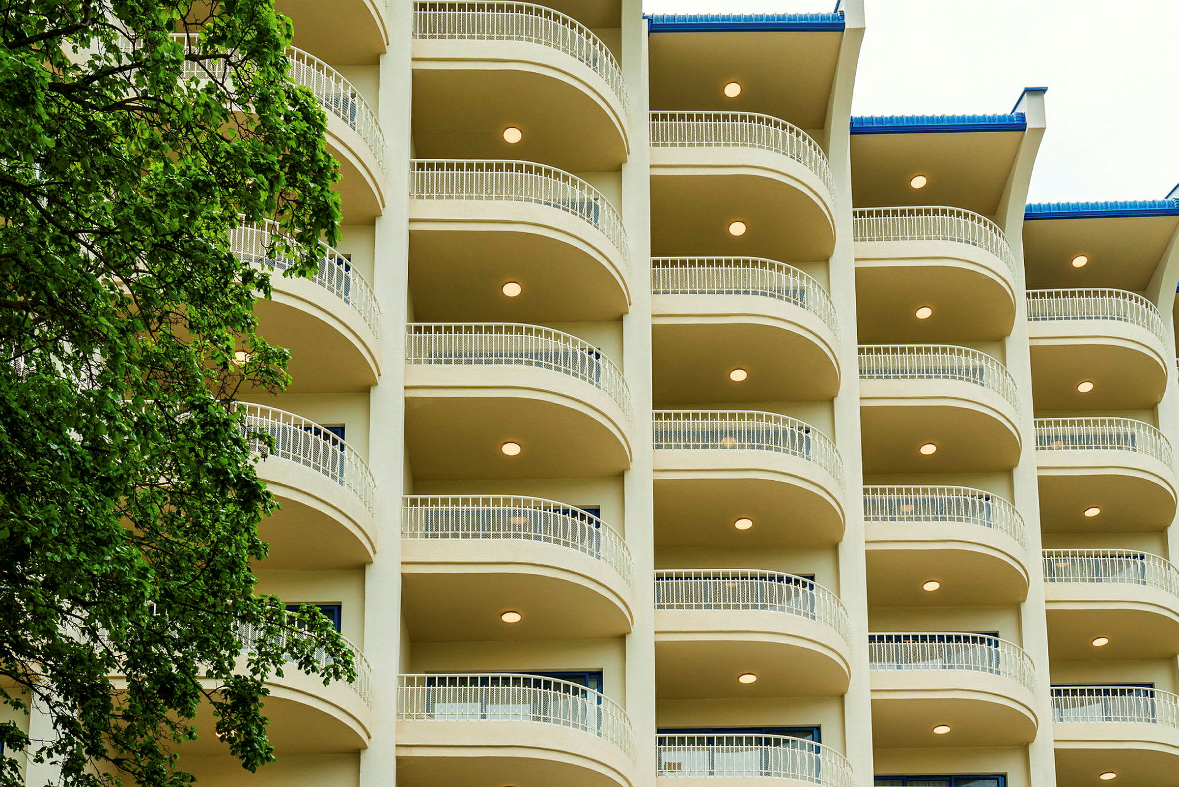 Facade af et hotel med flere etager og altaner. Et træ er placeret til venstre i billedet. (AI-genereret tekst)