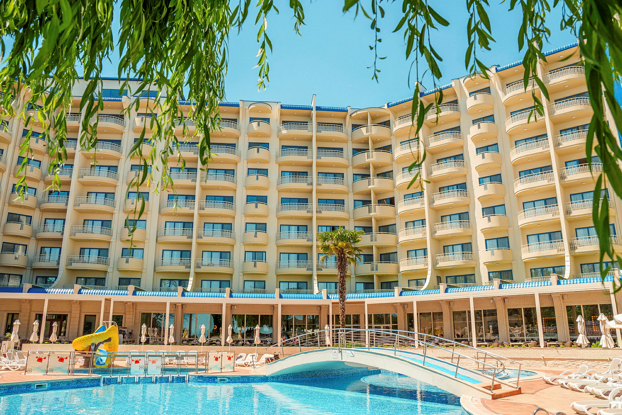 Hotellets facade med altaner, en pool med en bro og grønne planter i forgrunden. (AI-genereret tekst)