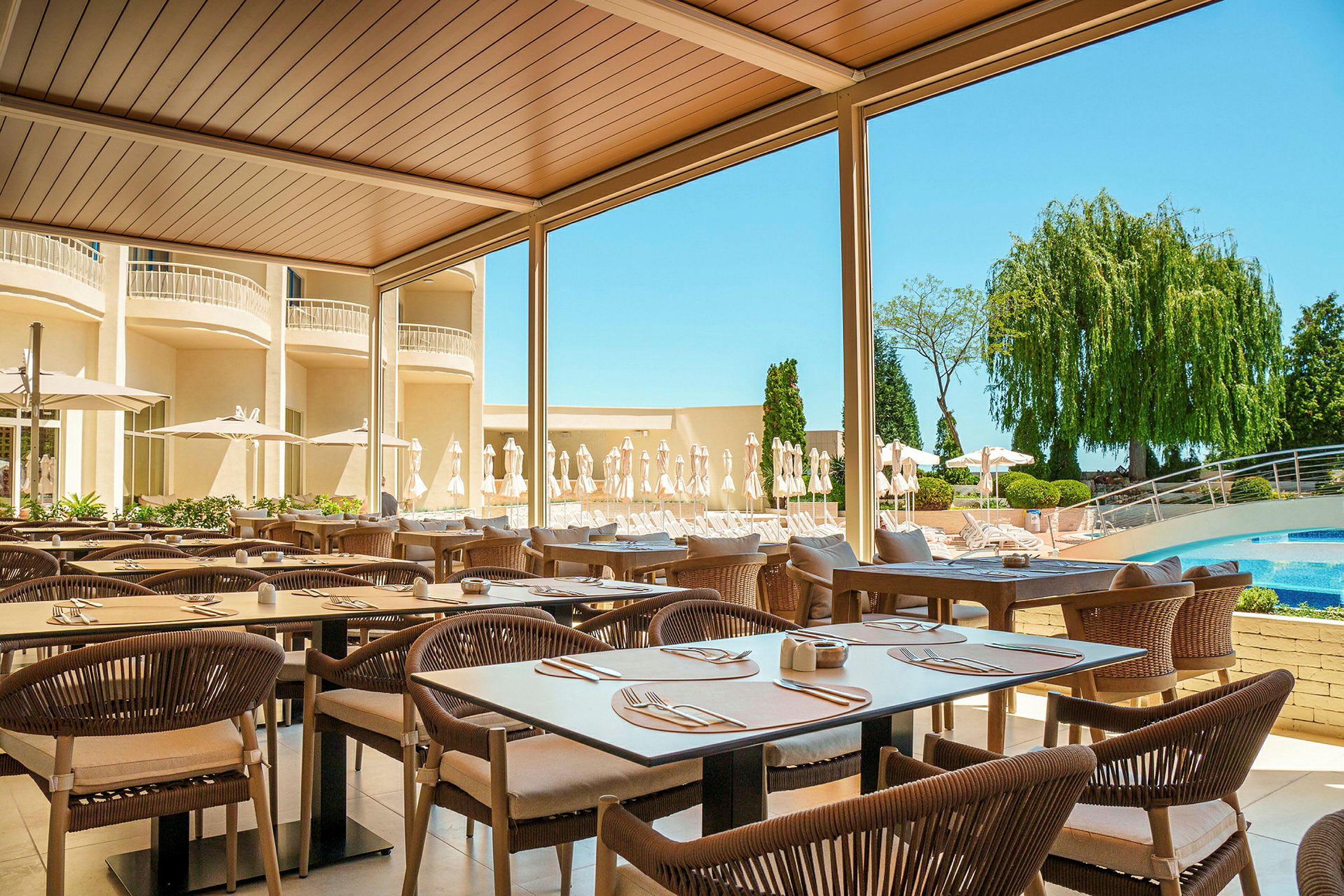 Udendørsrestaurant med borde og stole under tag, omgivet af grønt og en pool i baggrunden. (AI-genereret tekst)
