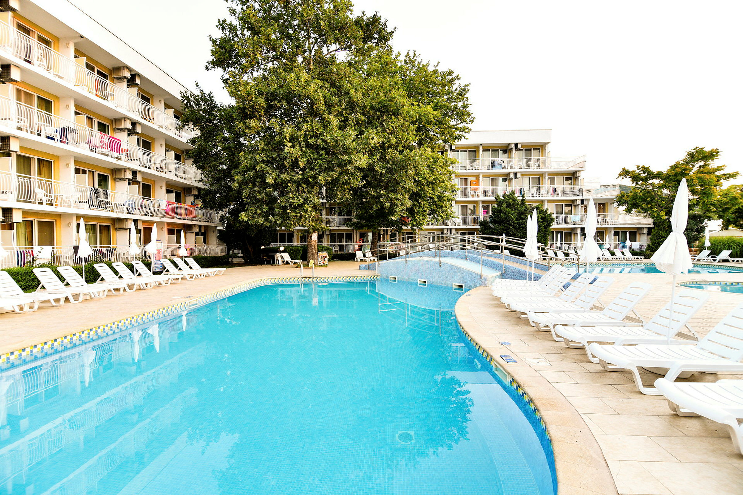 Billede af en udendørs pool ved et hotel med liggestole og beplantning. (AI-genereret tekst)