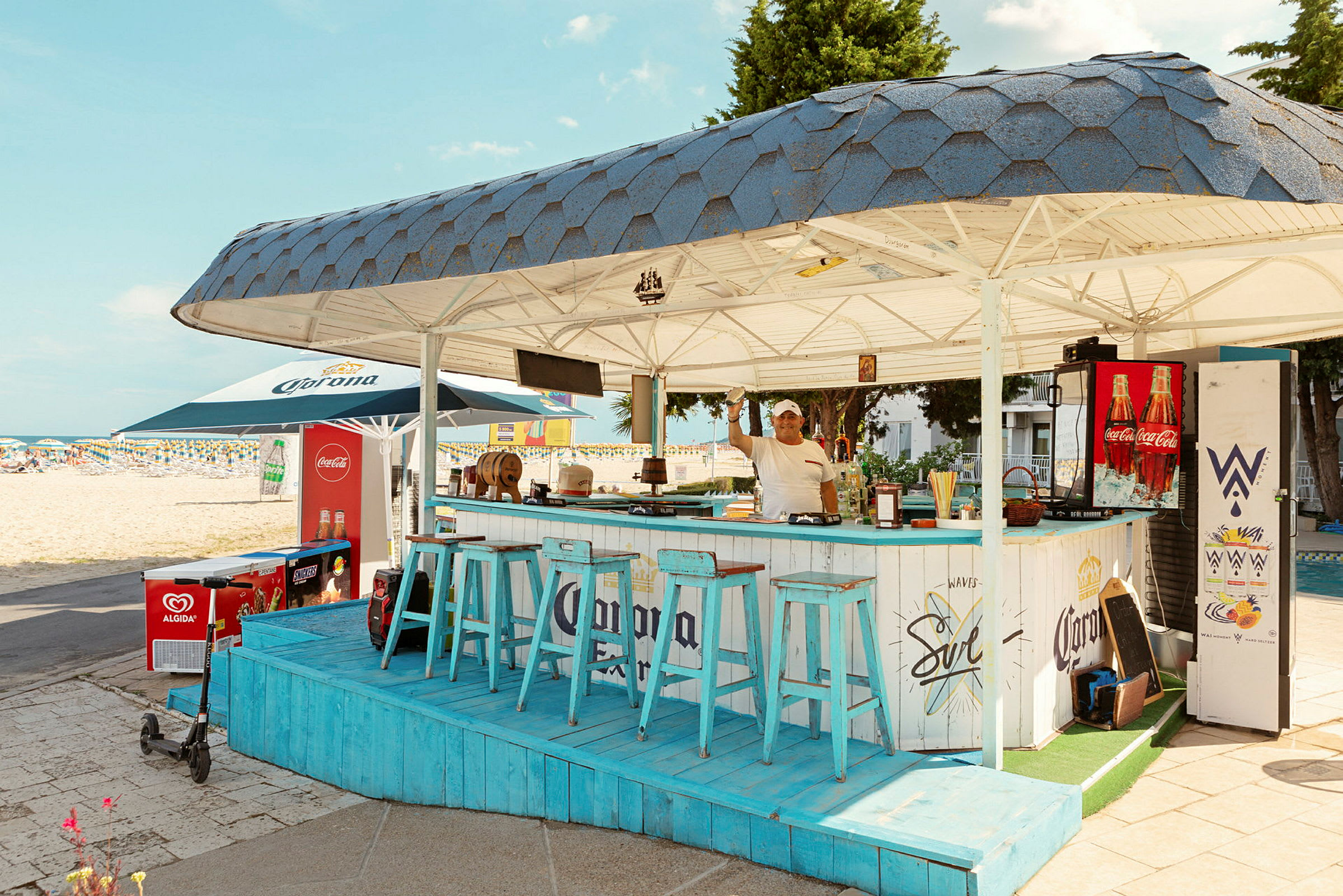 En strandbar med blå stole og tag, beliggende nær sandstranden. (AI-genereret tekst)