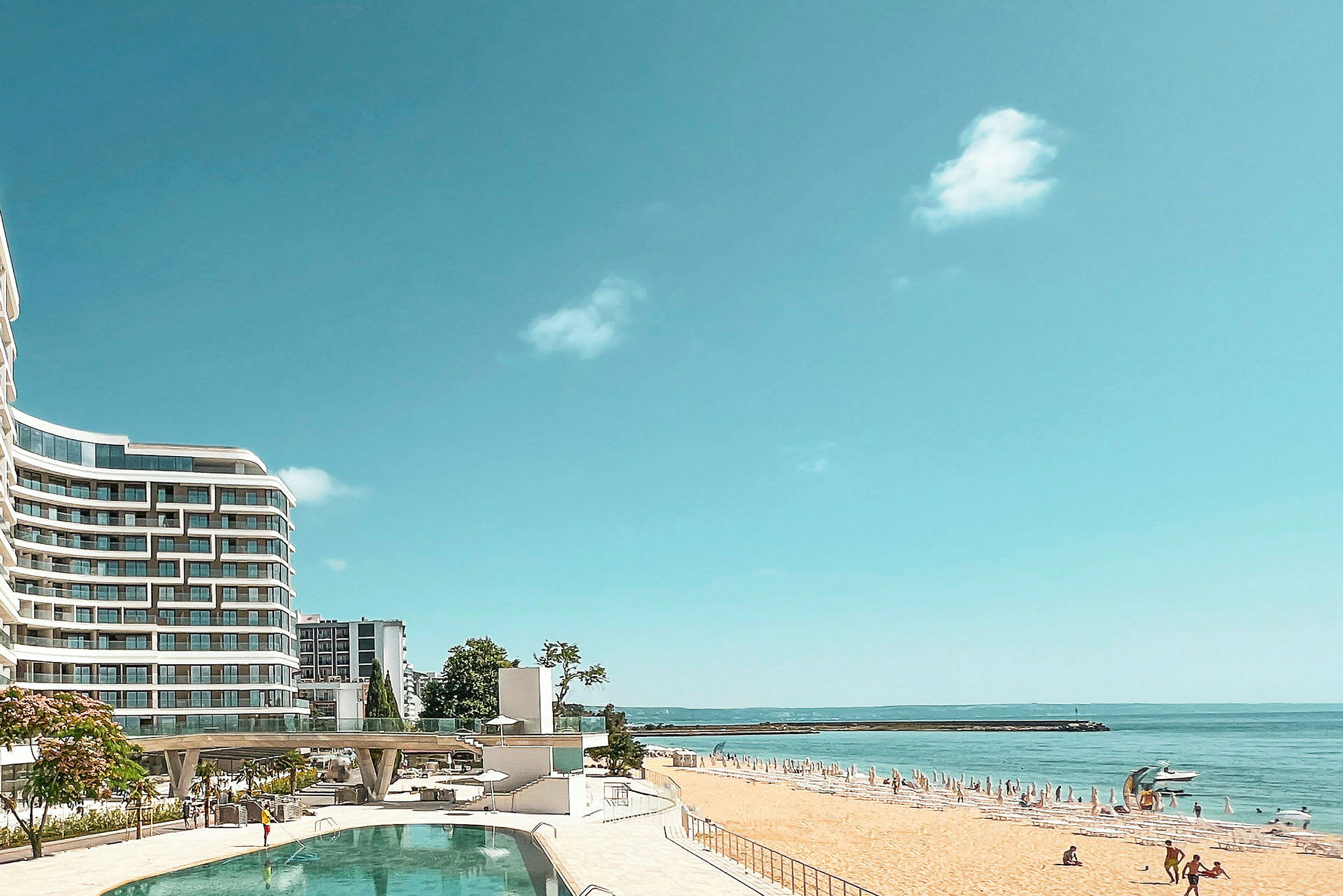 Hotelbygning ved en pool og en sandstrand med parasoller ved havet. (AI-genereret tekst)
