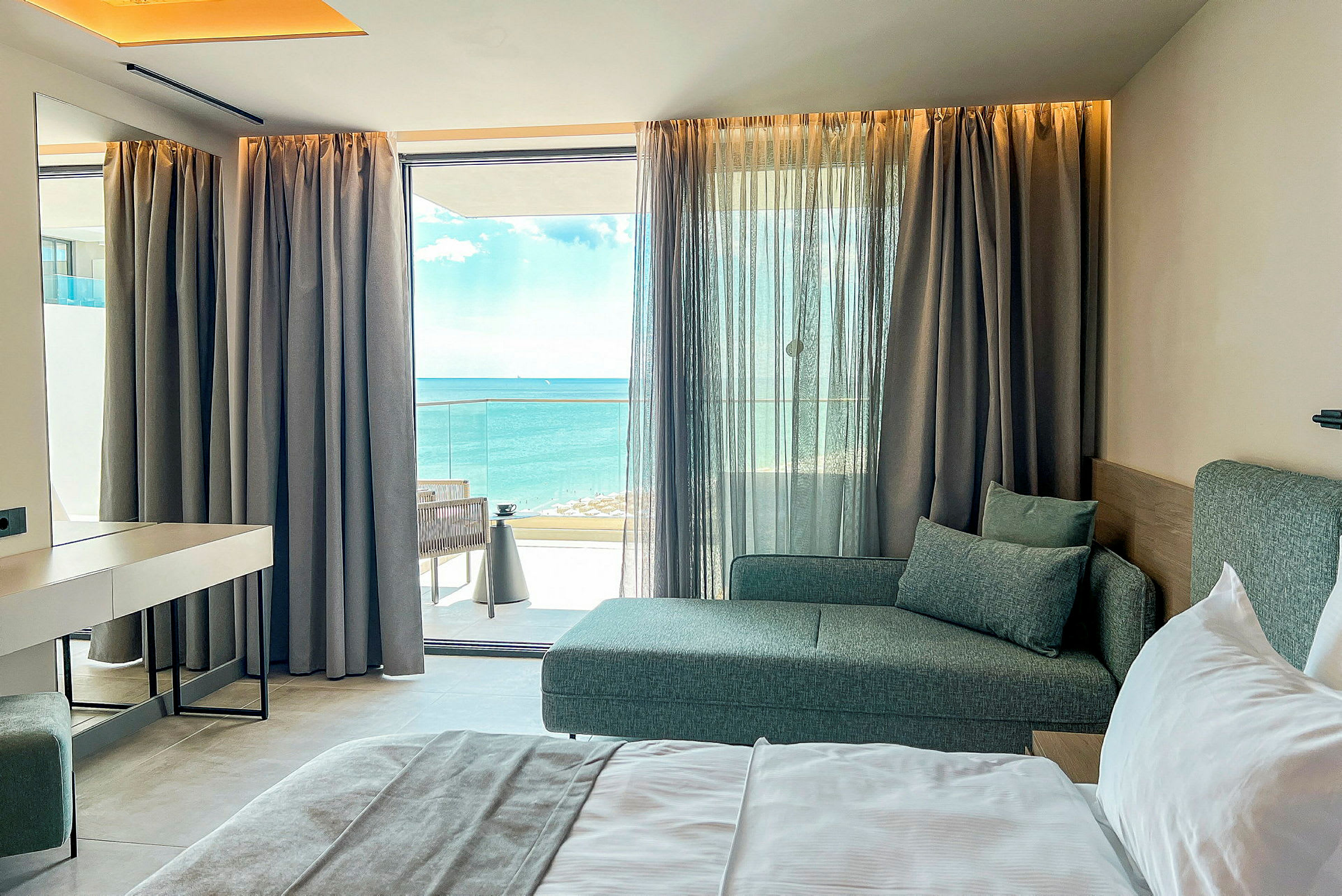 Hotelværelse med seng, sofa og havudsigt gennem en vinduesfront. (AI-genereret tekst)