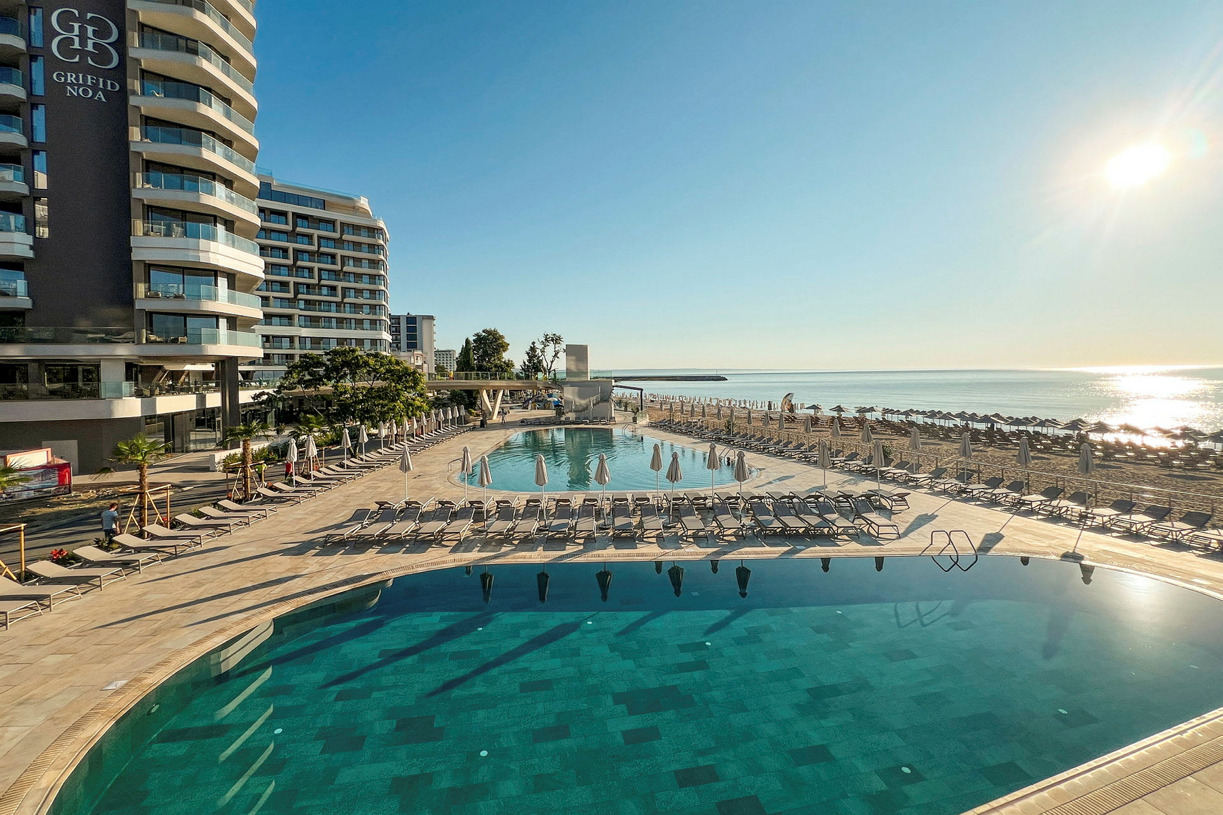 Hotelbygning ved strandpromenade med swimmingpool og liggestole. (AI-genereret tekst)