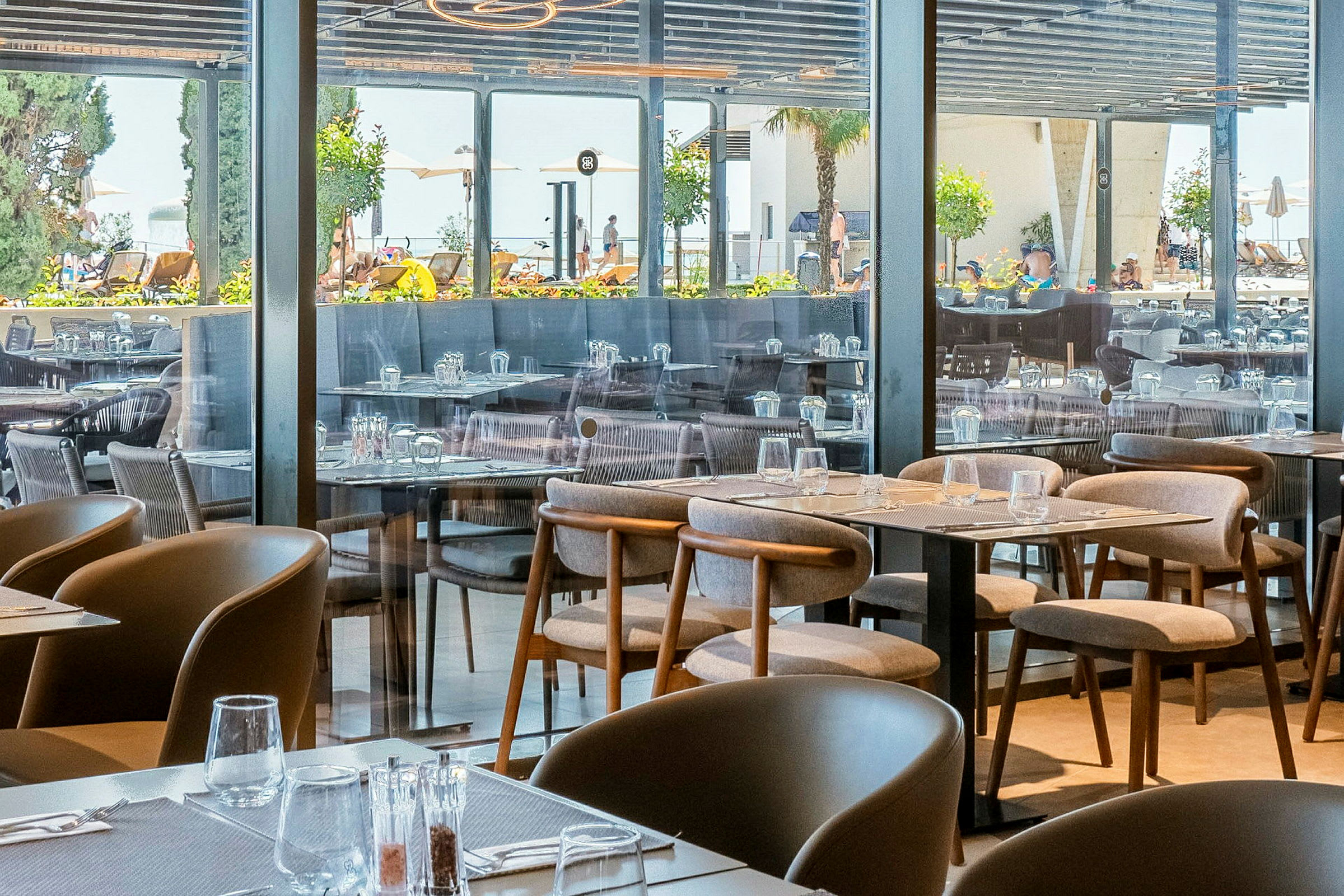 Billede af en restaurant med opstillede borde og stole, med udsigt mod udendørsområdet gennem glasvægge. (AI-genereret tekst)