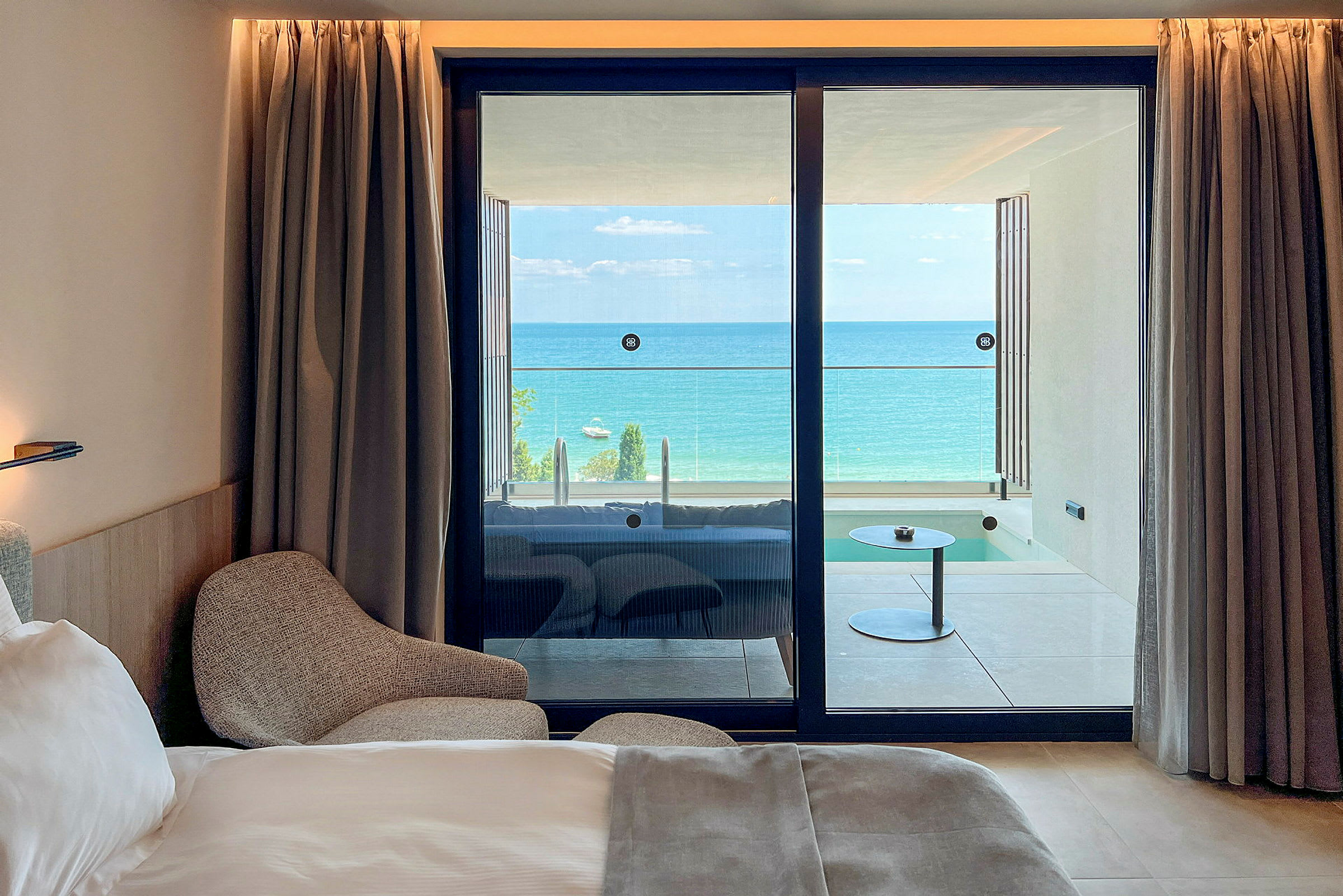 Et hotelværelse med en seng og lænestol, samt udsigt til en balkon med havet i baggrunden. (AI-genereret tekst)