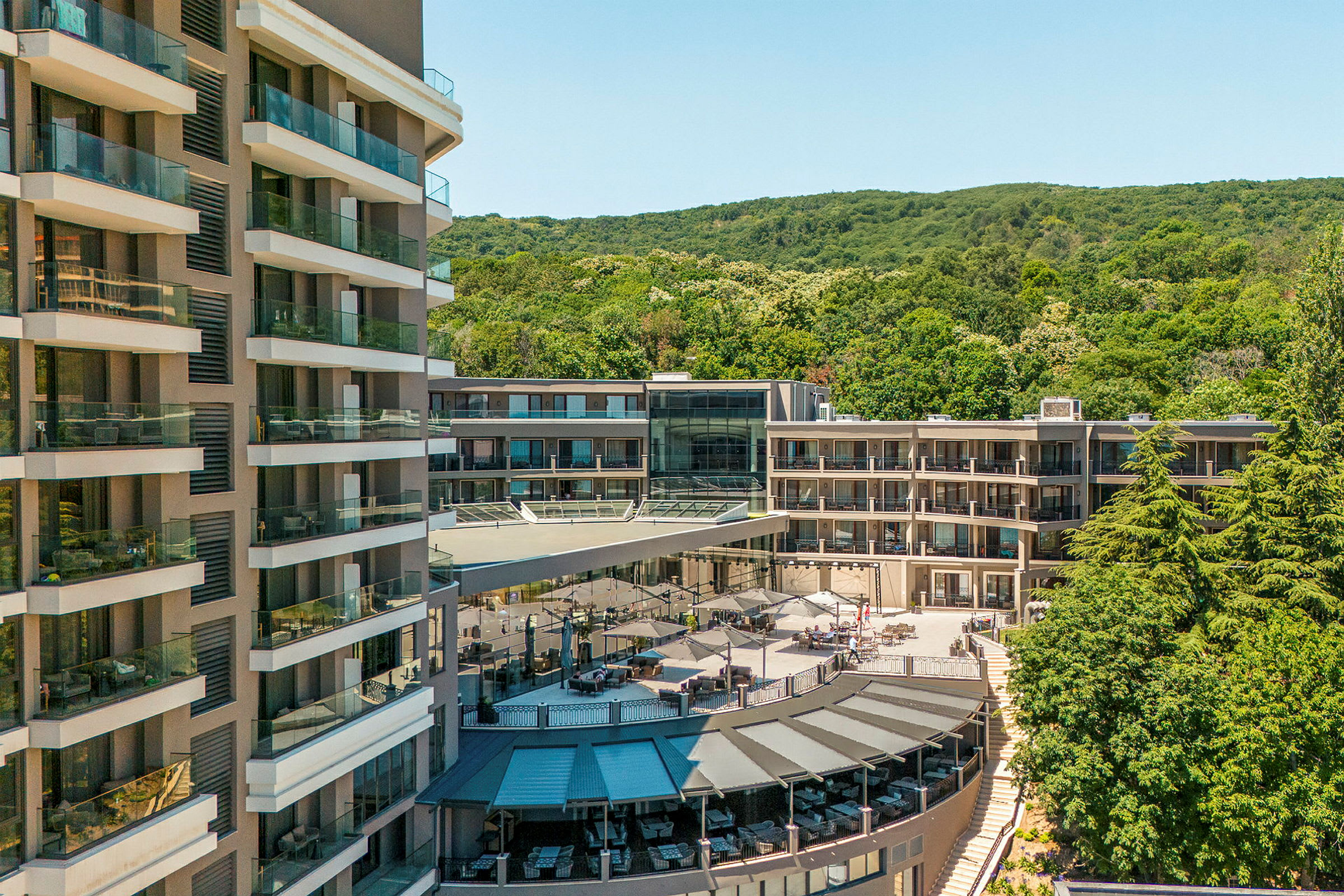 Flere hotelbygninger omgivet af grønt område og en terrasse med parasoller. (AI-genereret tekst)