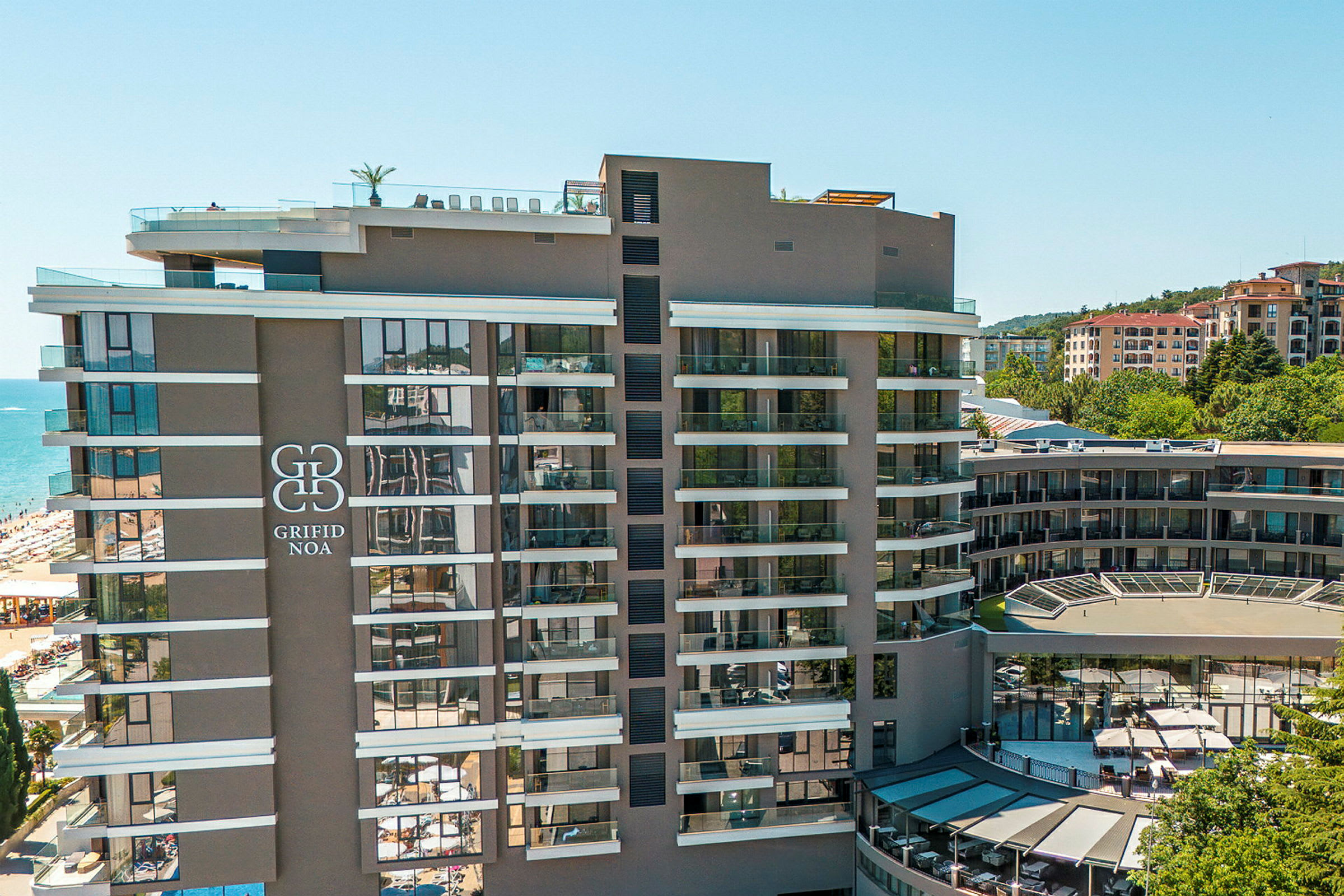 Moderne hotel med flere etager, stor logo på facaden, beliggende tæt på en strand og omgivet af bygninger. (AI-genereret tekst)